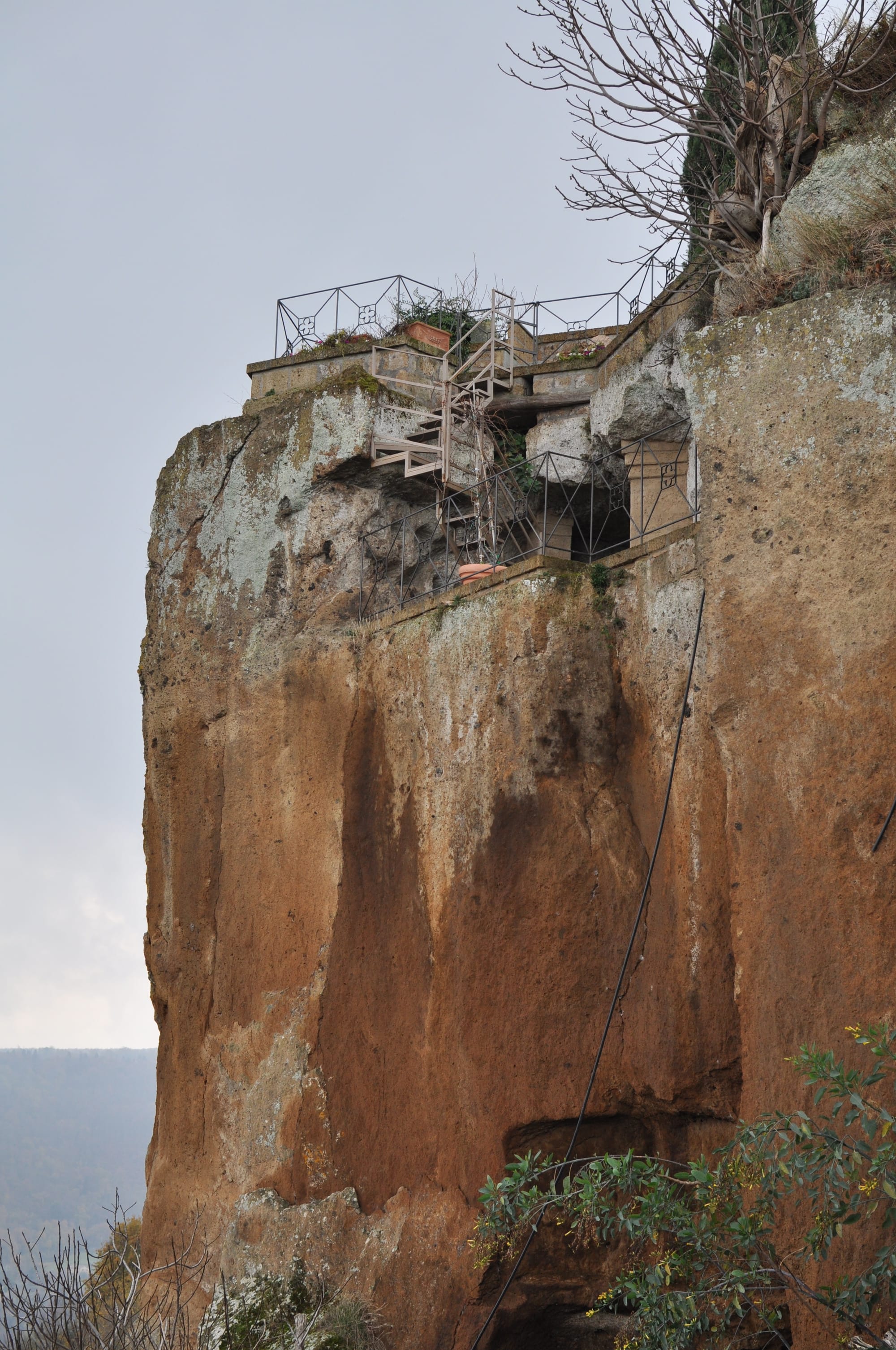 A vertical shot of a massive, sheer orange tufa cliff face topped with a stone terrace and metal railings under a grey sky.