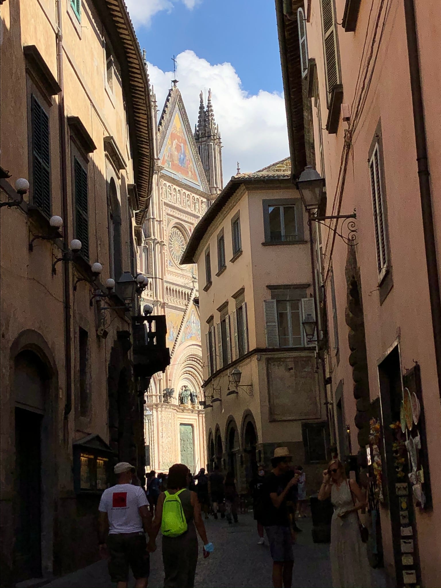 Low-angle view up a narrow medieval street in Orvieto, where the golden mosaics and towering spires of the Duomo facade emerge between old residential stone buildings.
