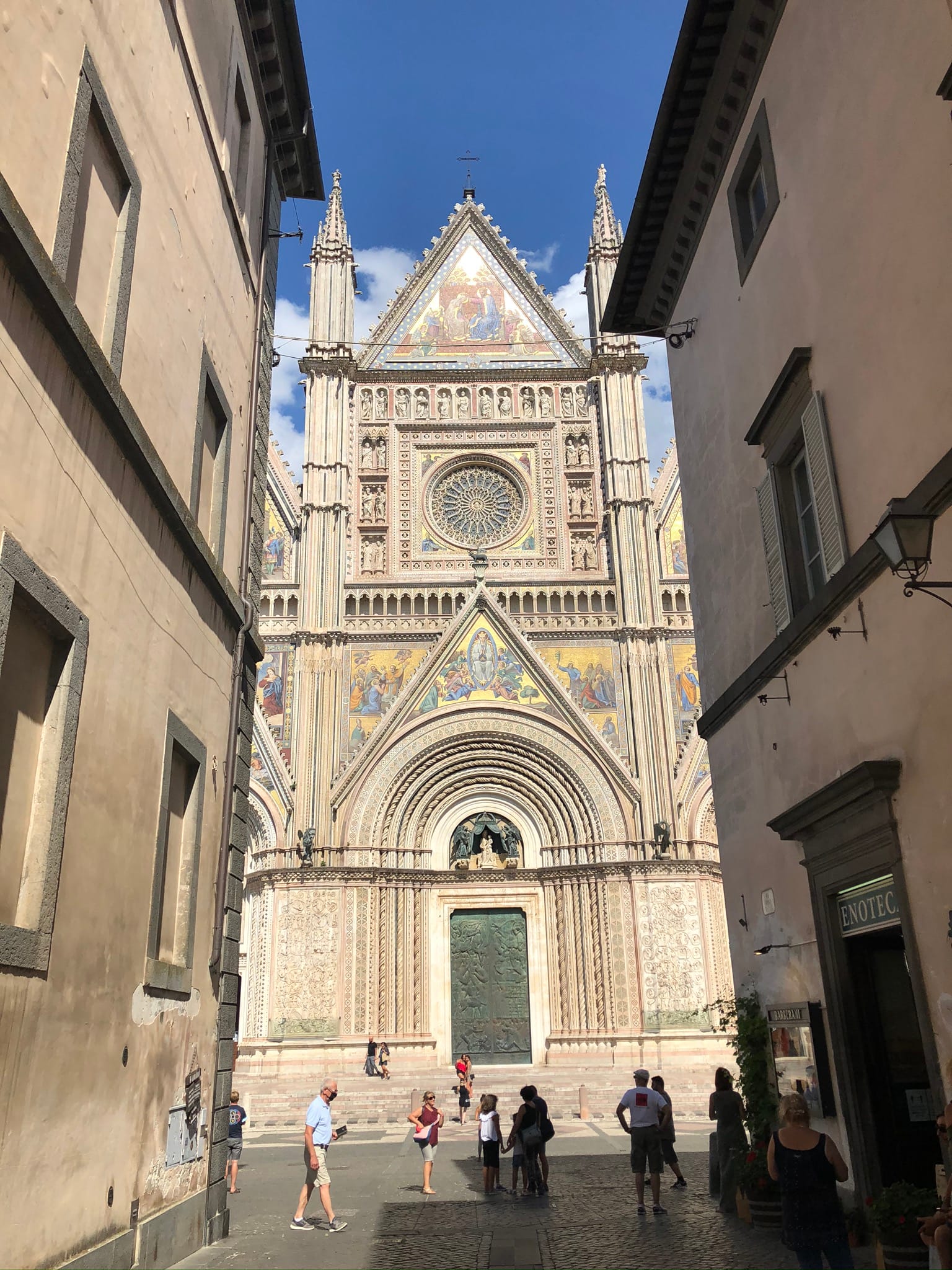 Symmetrical street-level view framed by historic buildings, looking down the axis towards the central portals and magnificent rose window of the Orvieto Duomo.