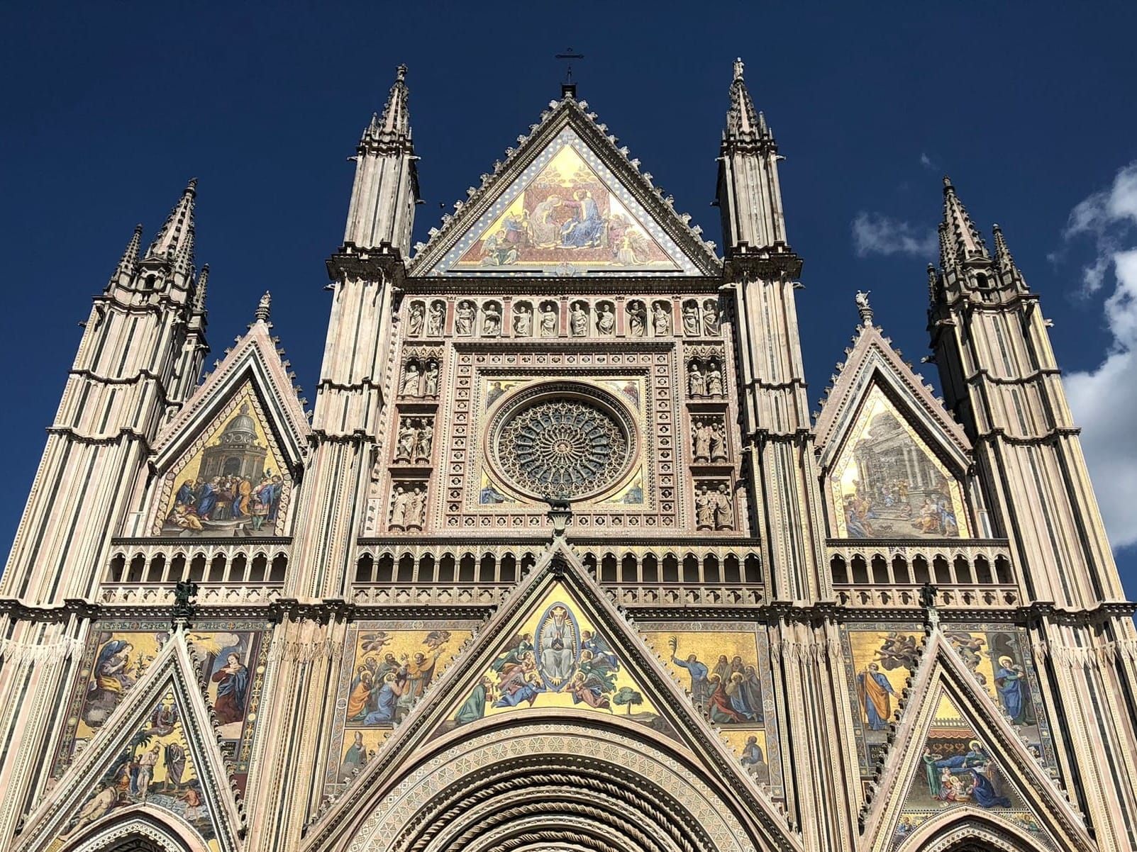 Frontal photograph of the Gothic facade of the Orvieto Duomo, showcasing its three-part division, central rose window, and golden mosaic spires against a blue sky.