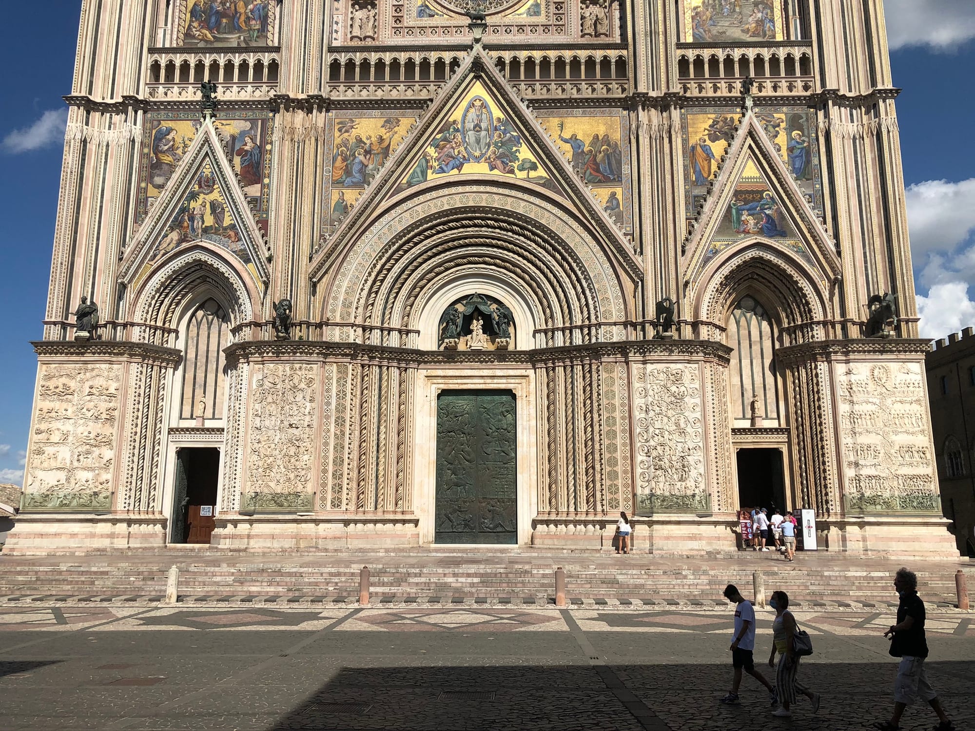 Lower half of the Duomo facade, showing the stone steps, three bronze doors, and heavily carved marble piers with biblical reliefs as visitors cross the piazza.