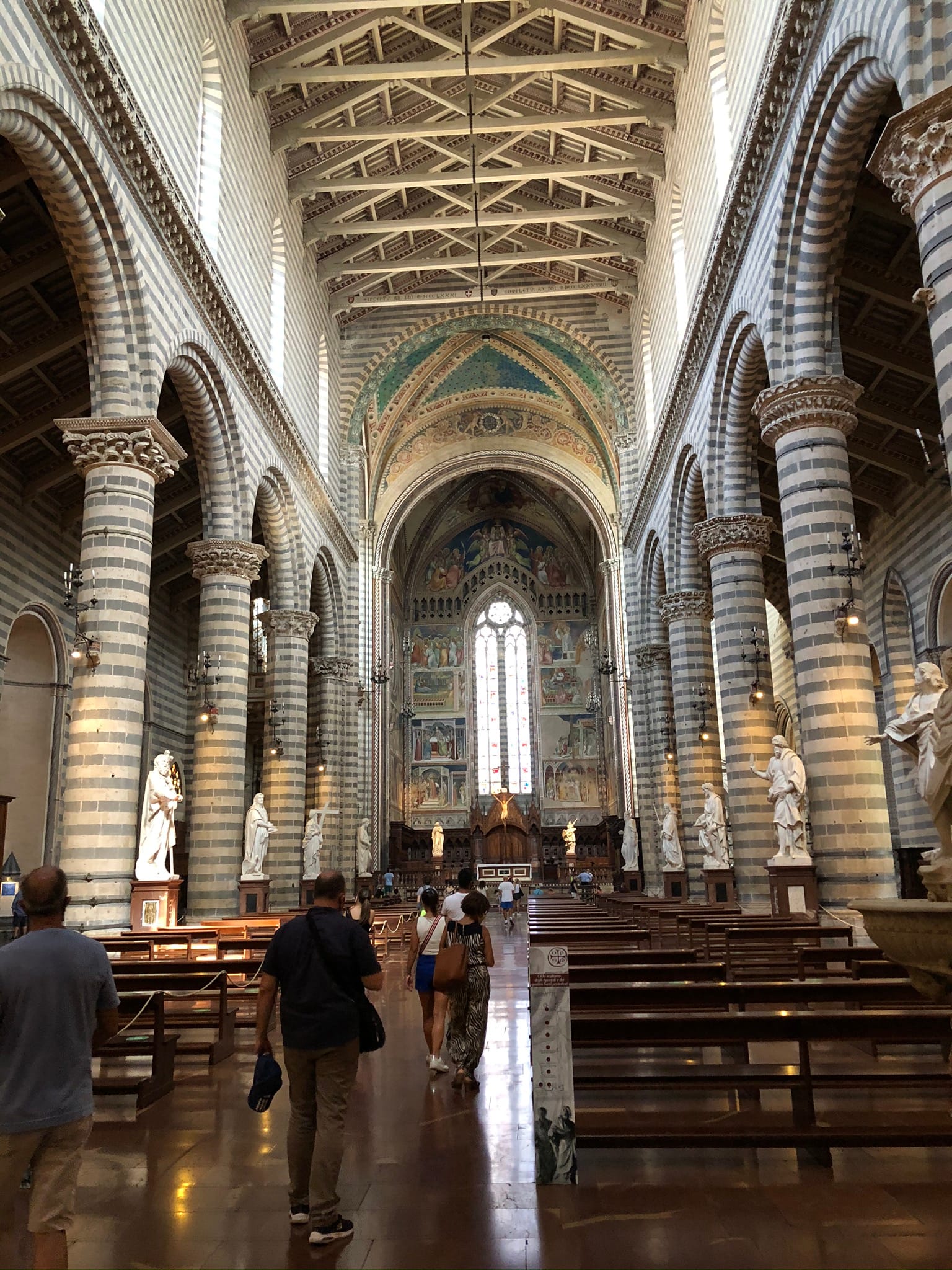 Interior view looking down the central nave toward the altar. Striped stone columns support round arches beneath an open wooden truss roof with marble statues.