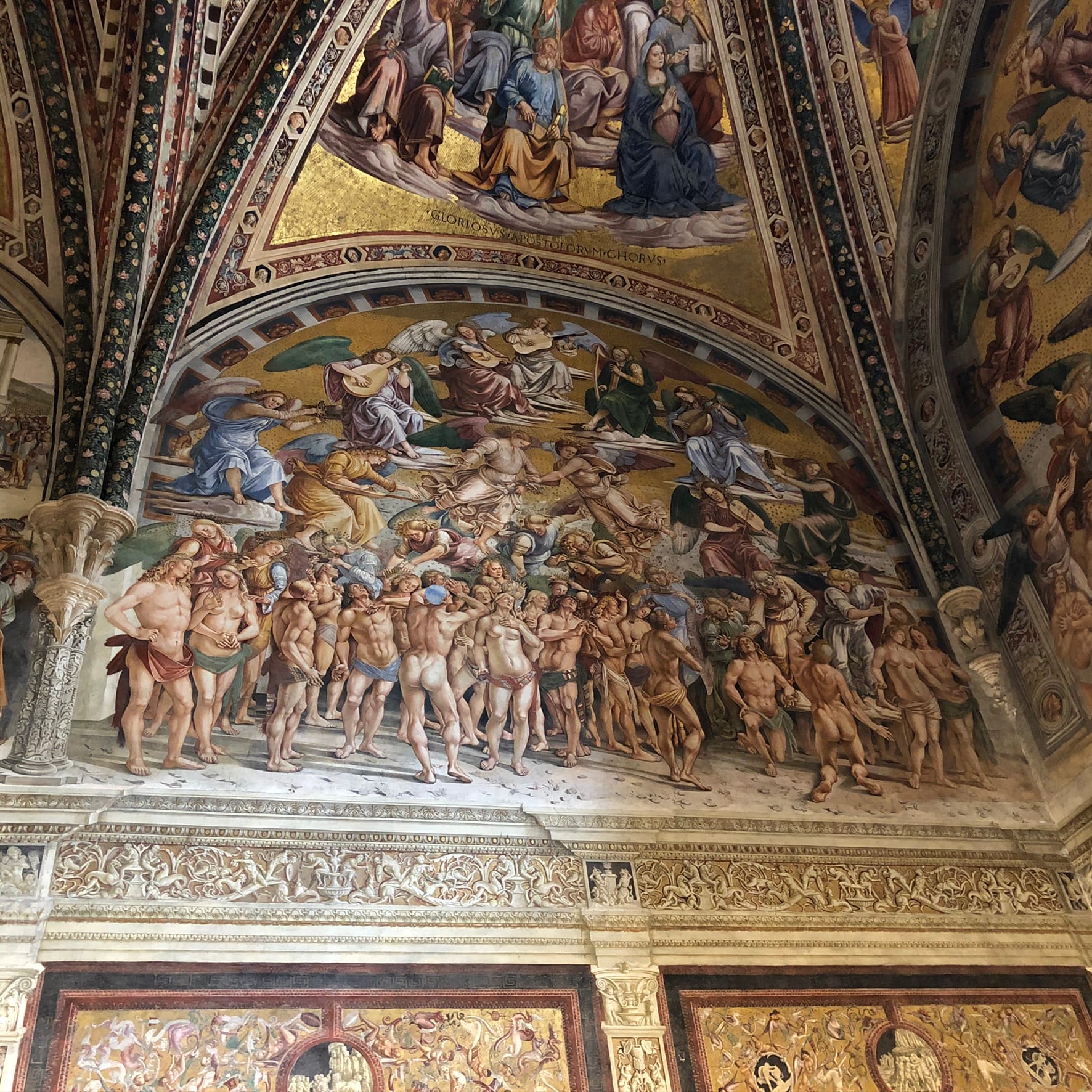 Renaissance fresco in the San Brizio Chapel showing a dense crowd of muscular nude figures below a sky filled with musical angels on golden clouds.