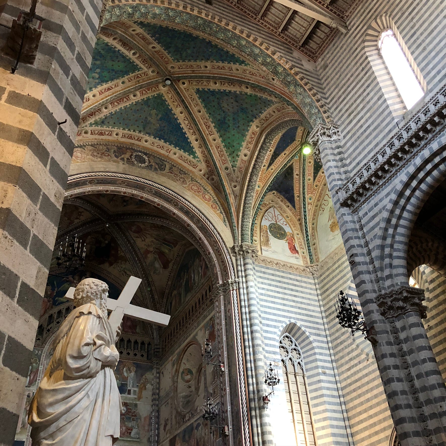 View from behind a marble statue looking up at alternating dark and light stone wall bands and a groin-vaulted ceiling with rich green and gold frescoes.