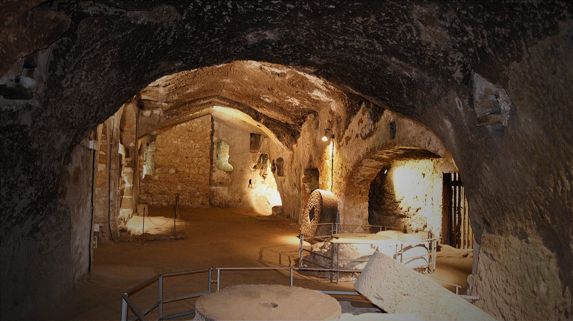 Dimly lit cavern in the Orvieto Underground, showing rock-carved arches, historic stone olive press millstones, and safety railings under a rugged tufa ceiling.
