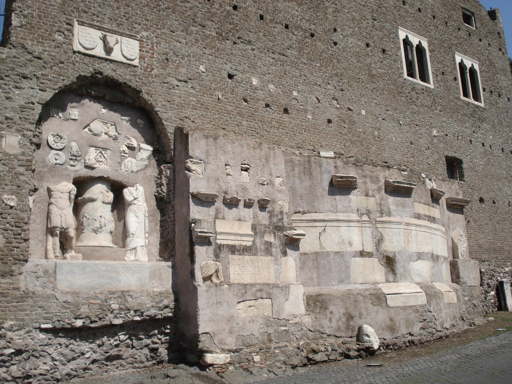 A brick wall with Gothic double windows above and, below, a niche with headless Roman statues and dozens of marble fragments, reliefs and inscriptions set into a pale plastered panel