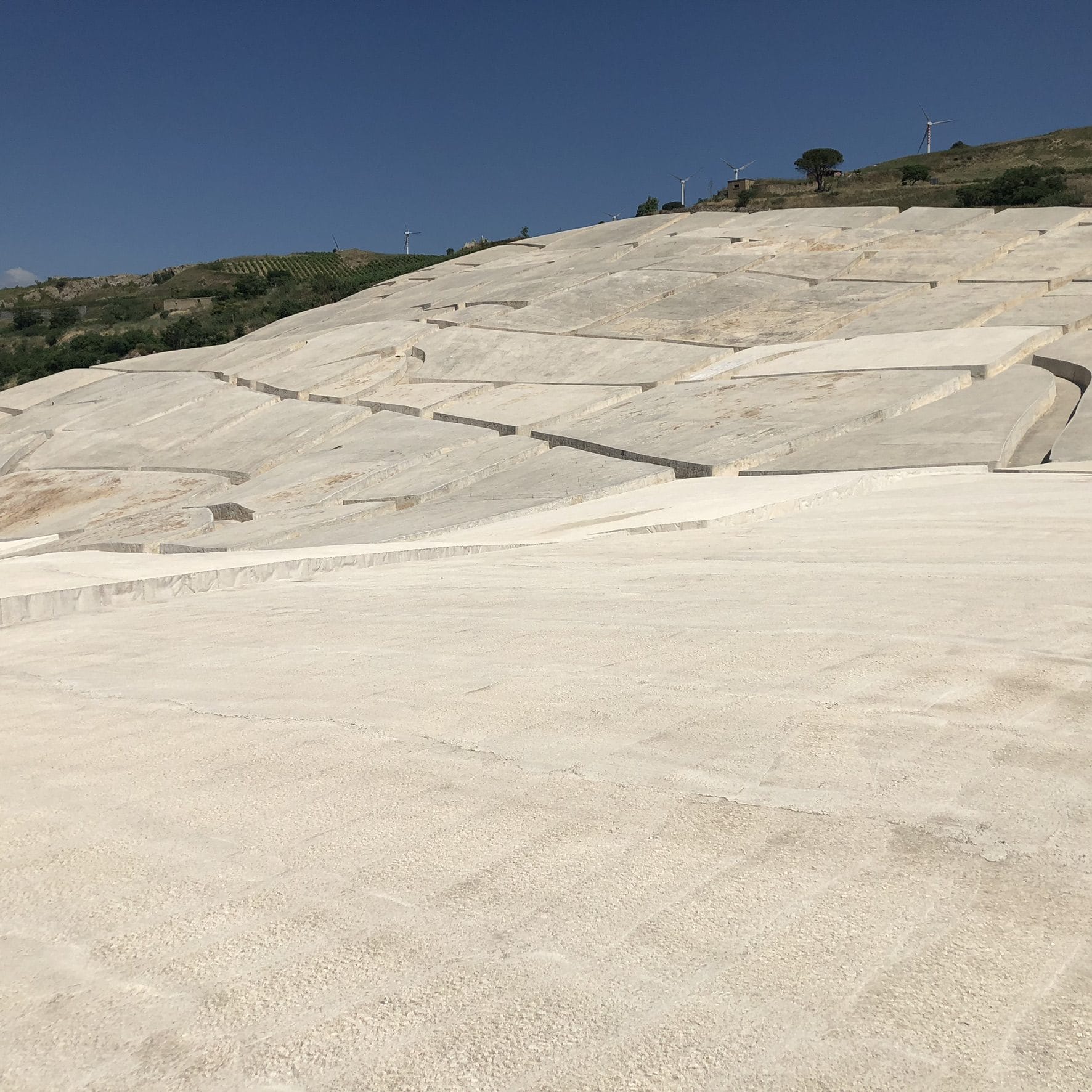 The white concrete blocks of Burri's Cretto spreading up a hillside under a blue sky, with wind turbines on the ridge above