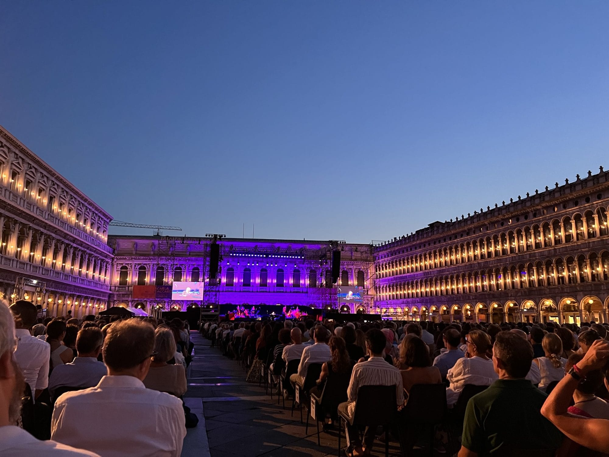 Audience seen from behind seated in Piazza San Marco at dusk, facing a stage lit in purple between the illuminated arcades of the Procuratie