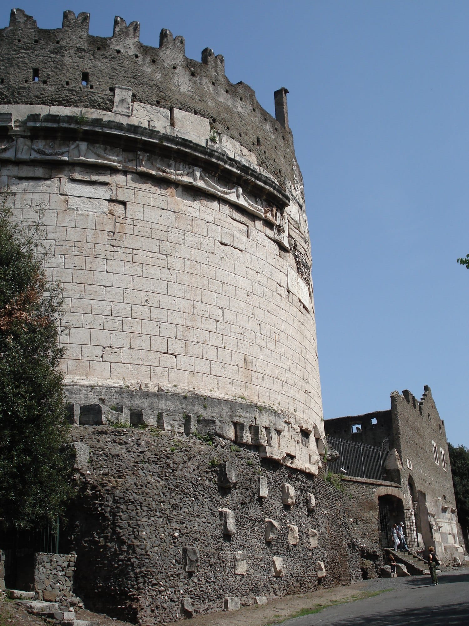 The travertine drum of the tomb of Caecilia Metella, crowned by medieval battlements, standing on a dark stone base beside a road that slopes away 
