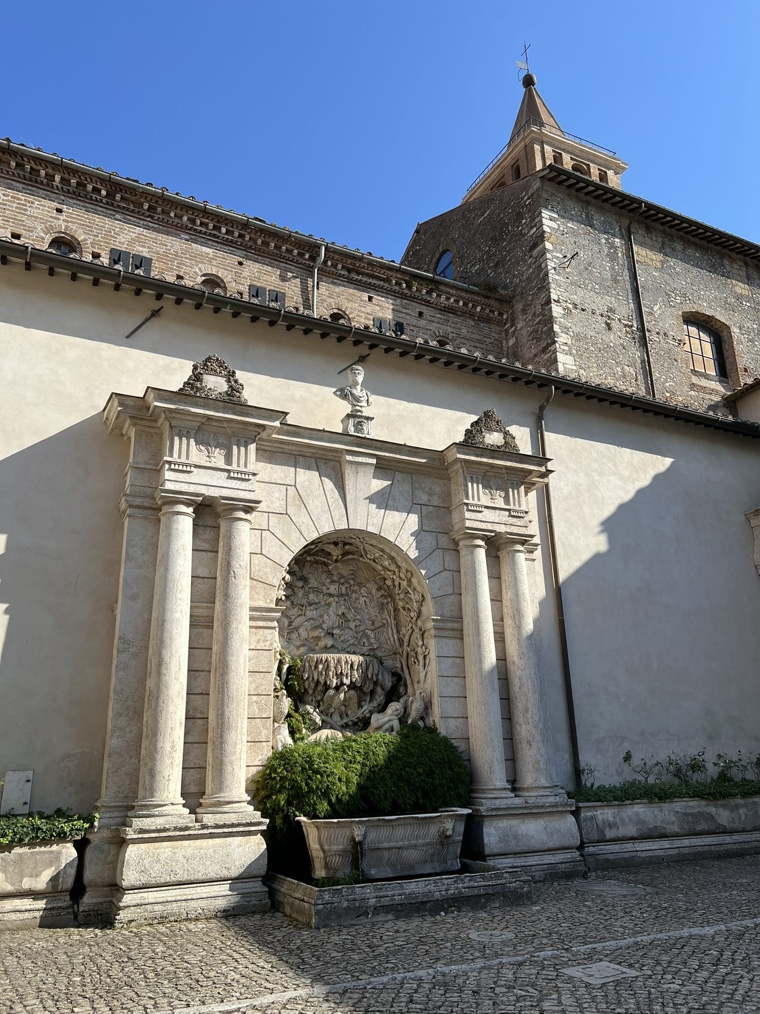 Fountain in a niche framed by columns and a pediment, on one side of the courtyard, with the bell tower behind