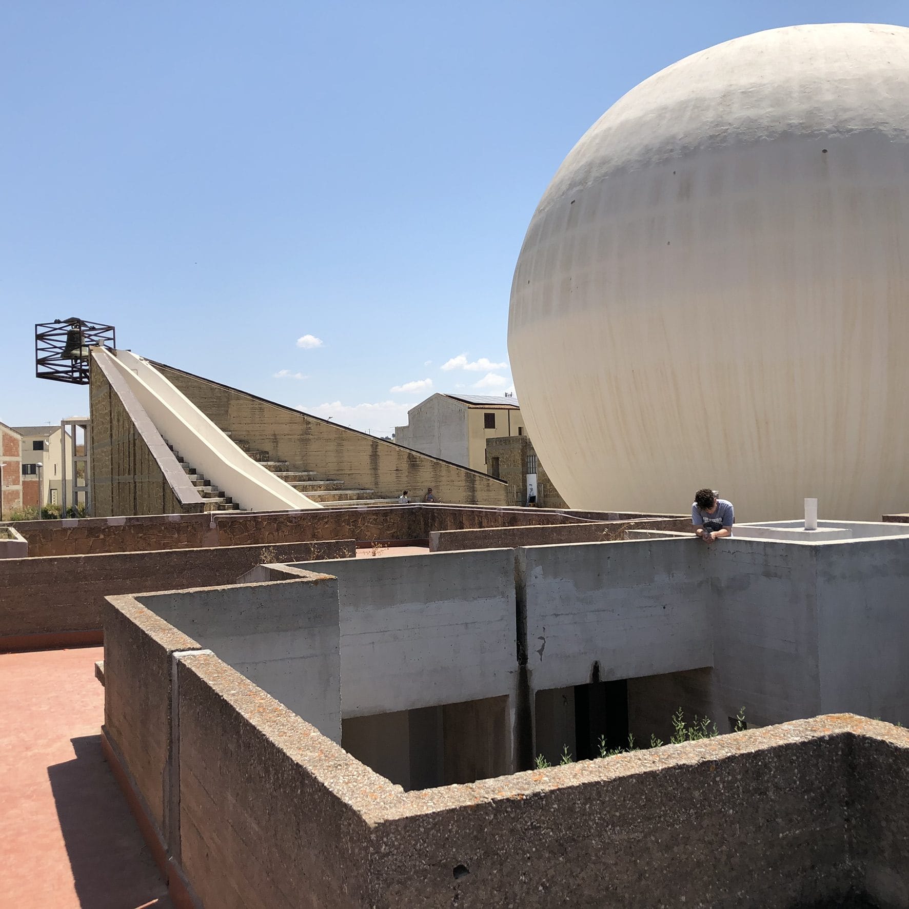 A large white sphere set within the stone and concrete walls of the Chiesa Madre of Gibellina Nuova, with a sloping bell tower behind
