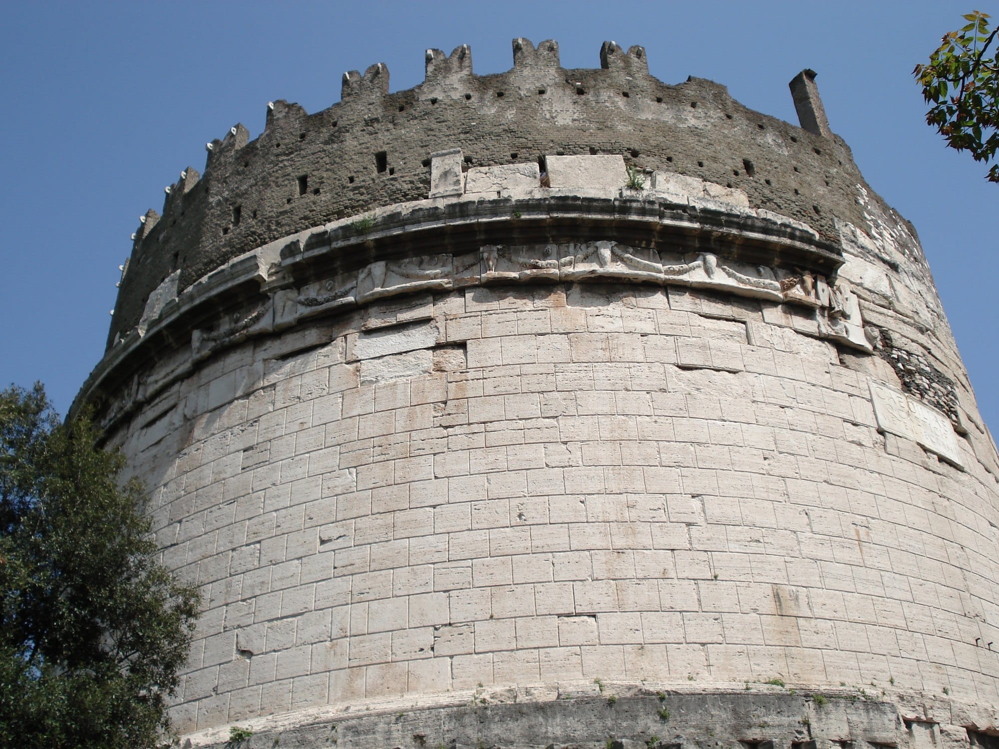 Close view of the curved travertine drum of the tomb of Caecilia Metella, with a carved frieze near the top and a rough medieval wall with battlements above it, against a blue sky 