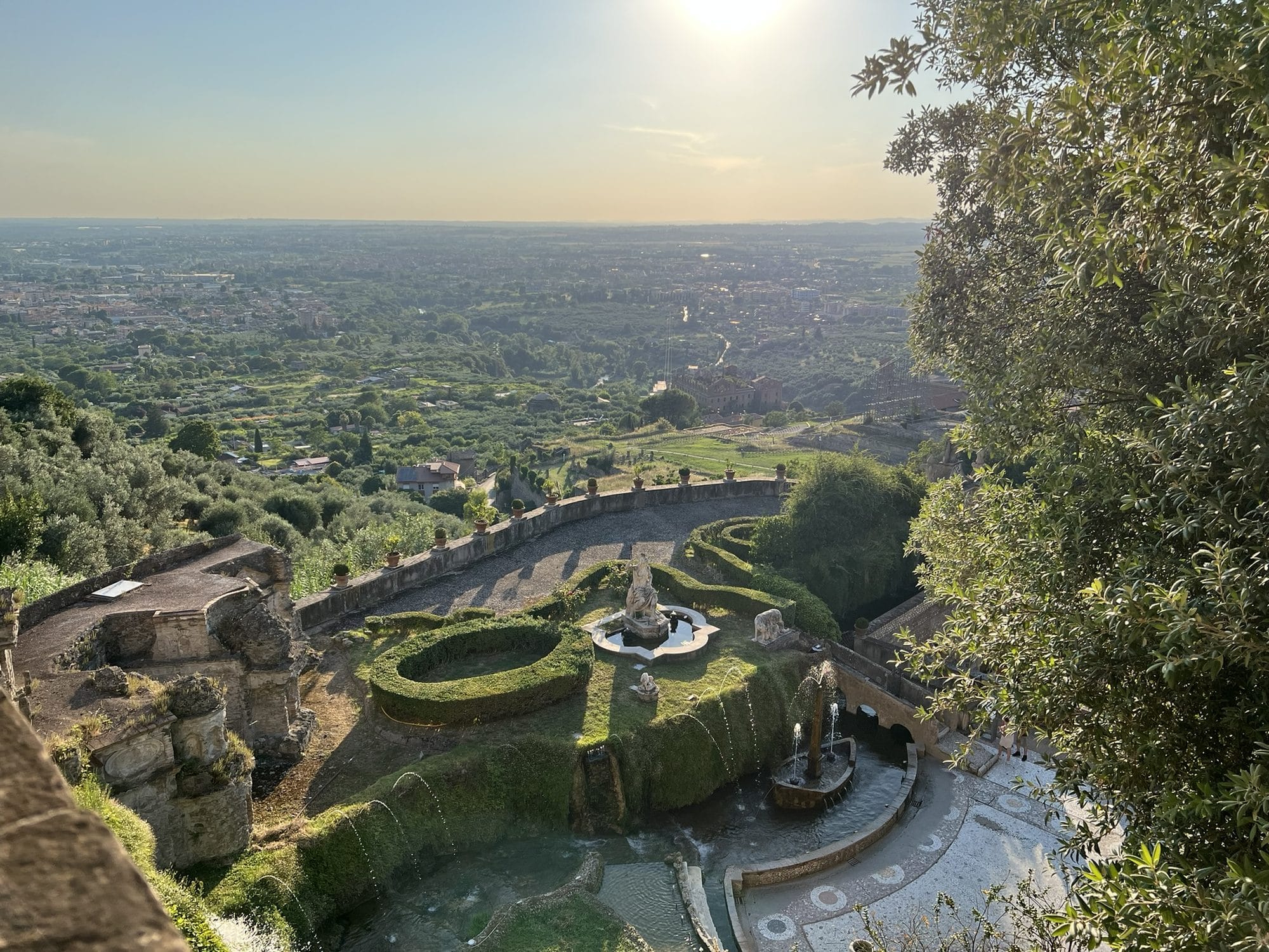 View from above of the Rometta fountain with statues, artificial rocks and hedges, and the plain in the golden evening light