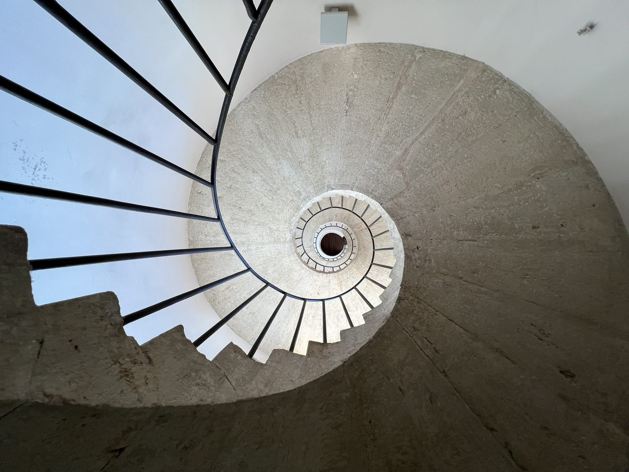 Spiral staircase in pale stone seen from below, its dark railing drawing a spiral