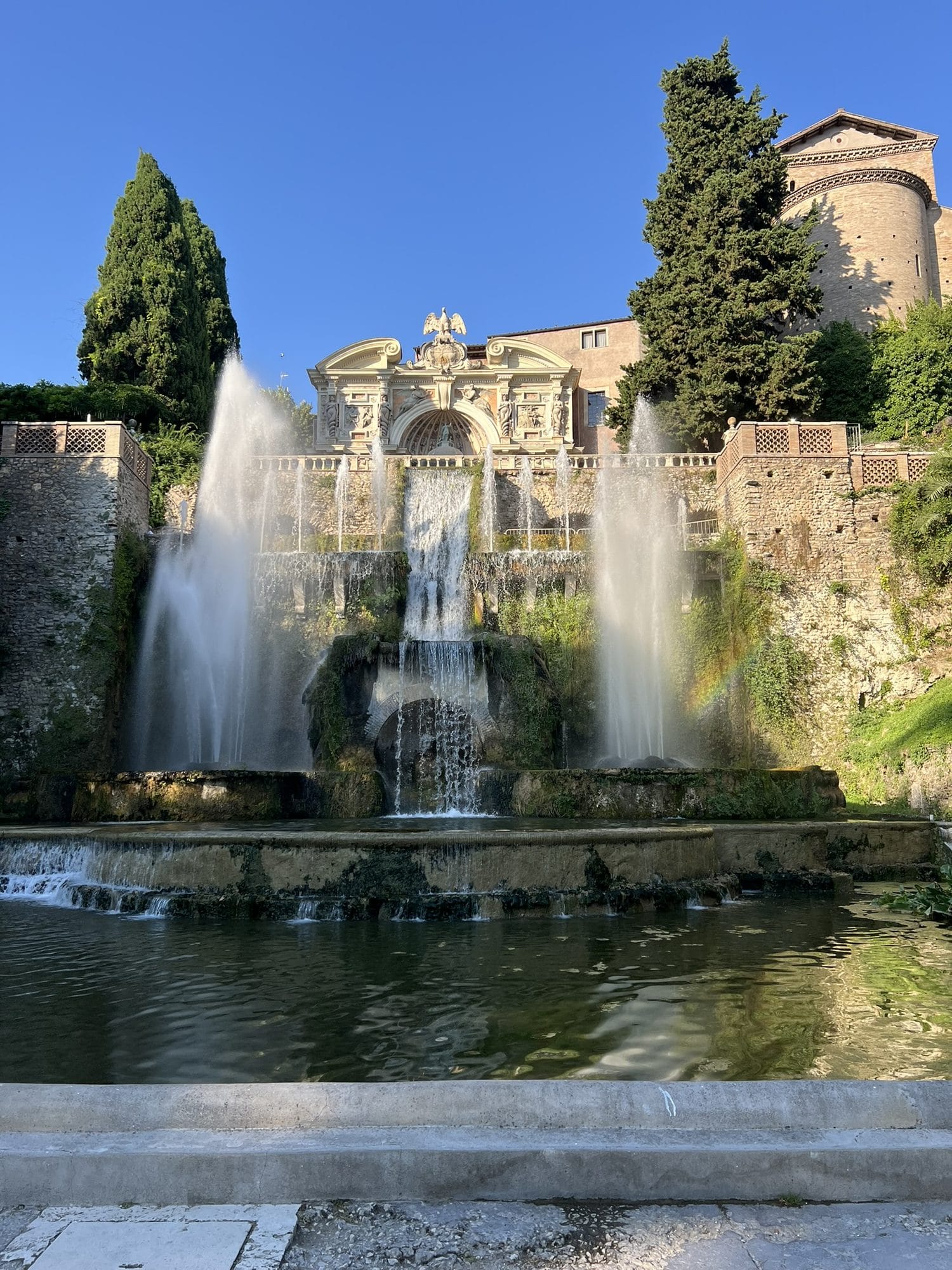 Cascades and tall jets of the Fountain of Neptune with a small rainbow on the right, under a blue sky