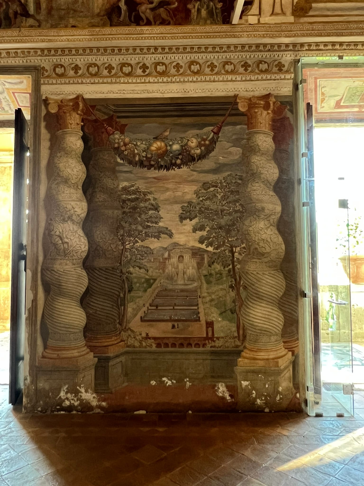Frescoed wall with two painted twisted columns framing a view of a terraced garden with fountains and a building at the top