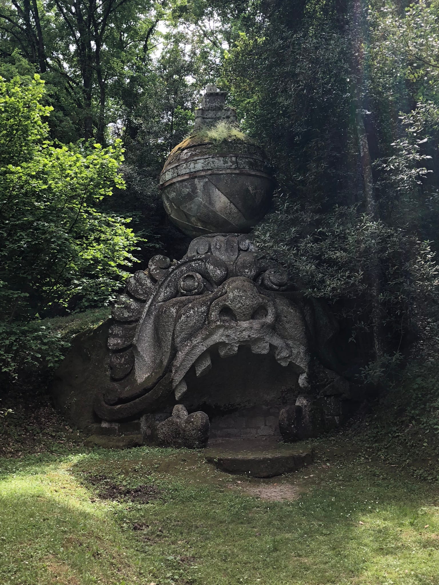 Colossal stone mask carved into the hillside with wide open jaws, supporting a stone sphere topped with a miniature castle, surrounded by trees at Bomarzo.