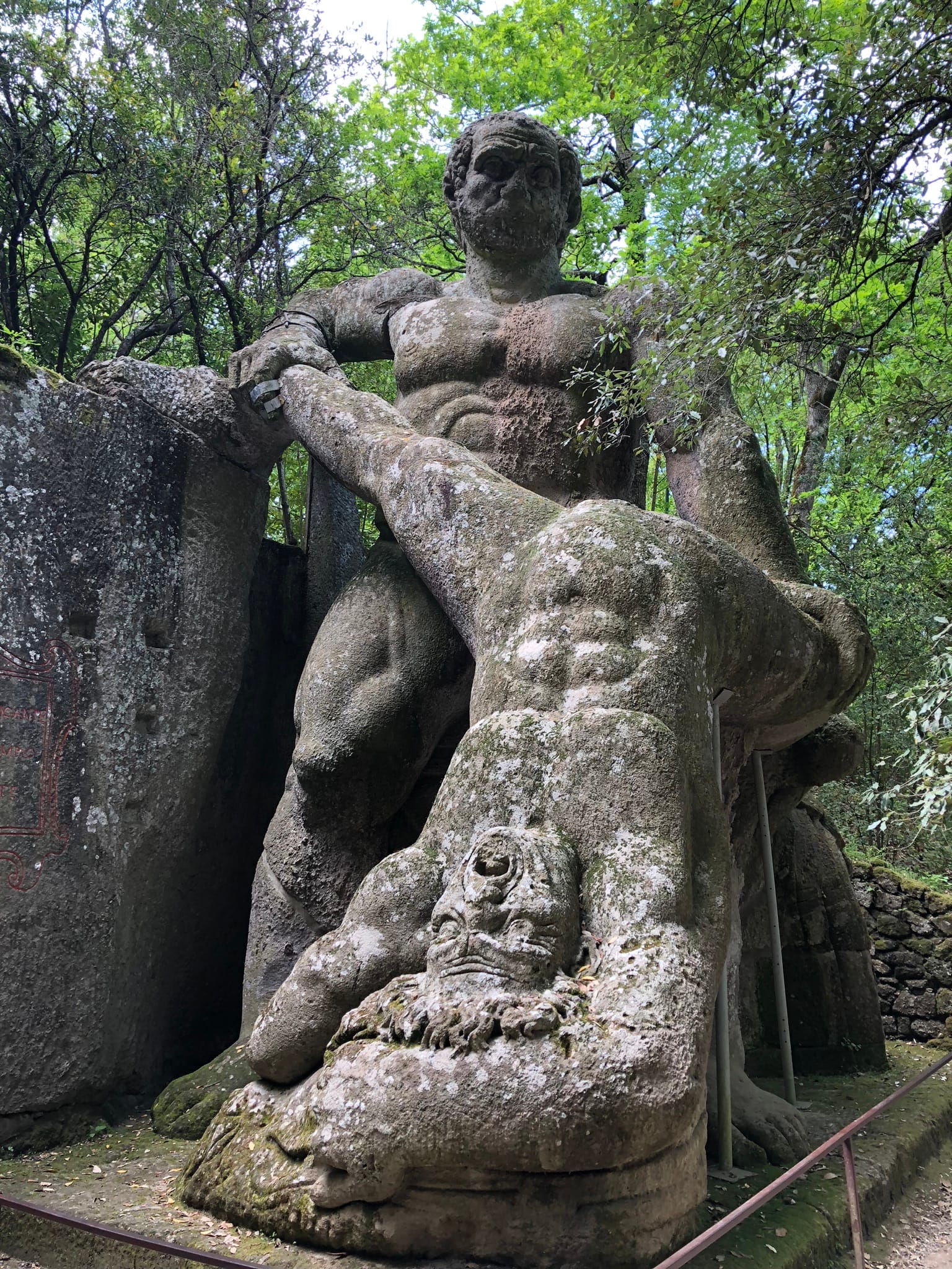 Colossal stone sculpture of Hercules holding the defeated giant Cacus upside down by his legs, surrounded by trees in the Sacro Bosco of Bomarzo.