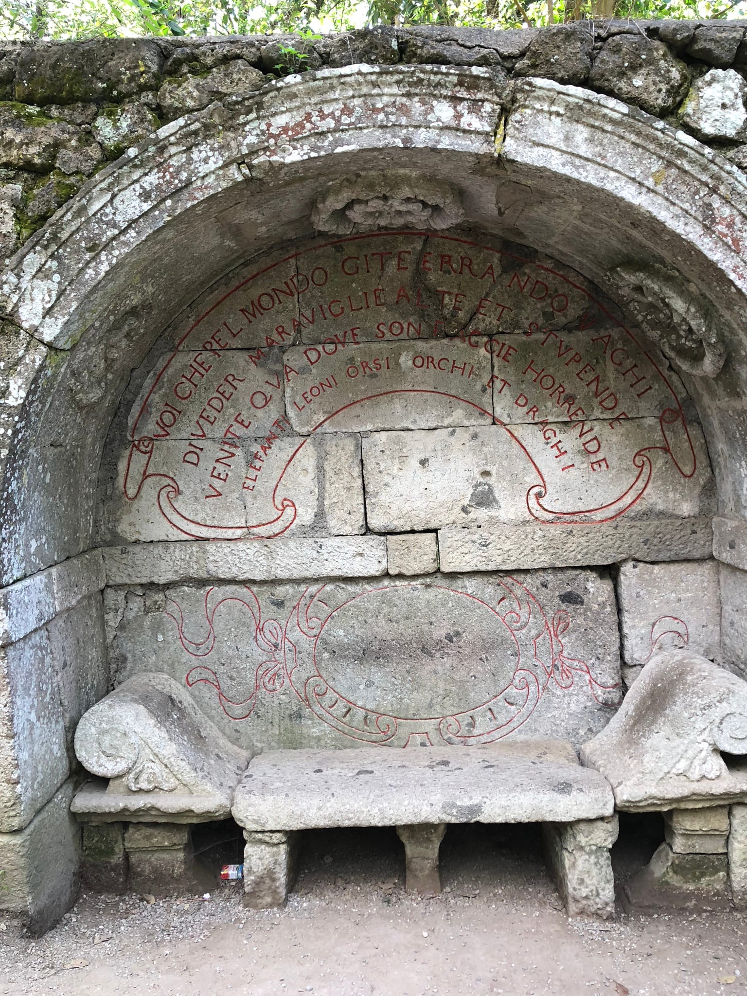 A weathered stone bench resembling an ancient Roman triclinium, carved inside a large vaulted natural niche in a moss-grown peperino boulder, surrounded by forest undergrowth and fallen leaves in the Sacro Bosco of Bomarzo.