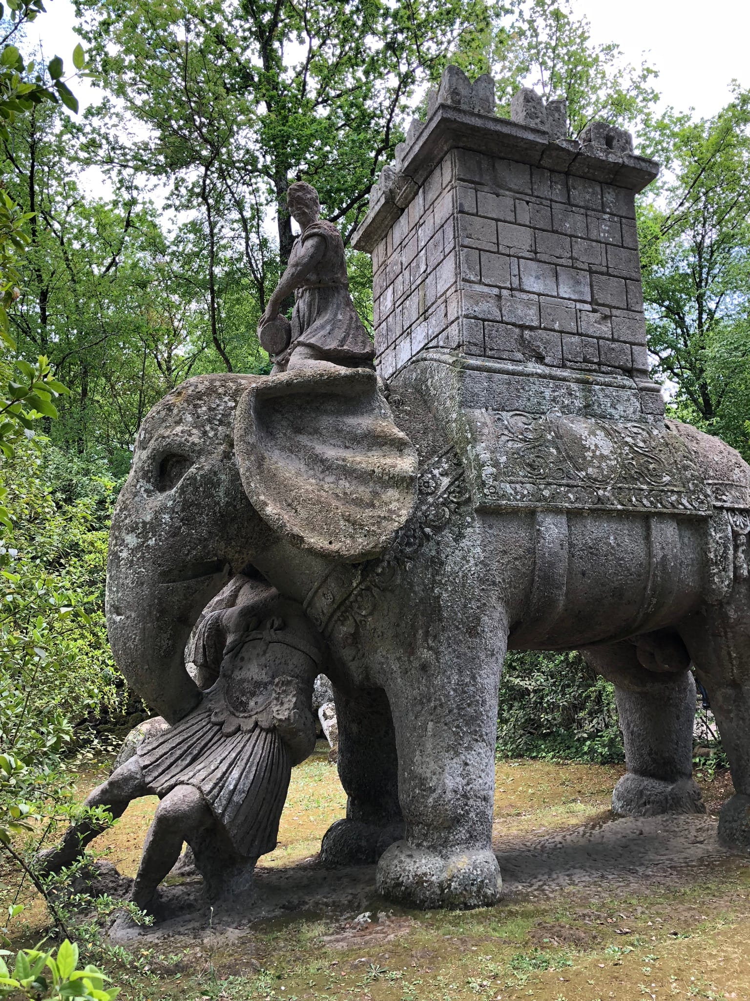 Colossal stone sculpture of an elephant carrying a fortified tower and holding a Roman soldier in its trunk, among the trees of Bomarzo.