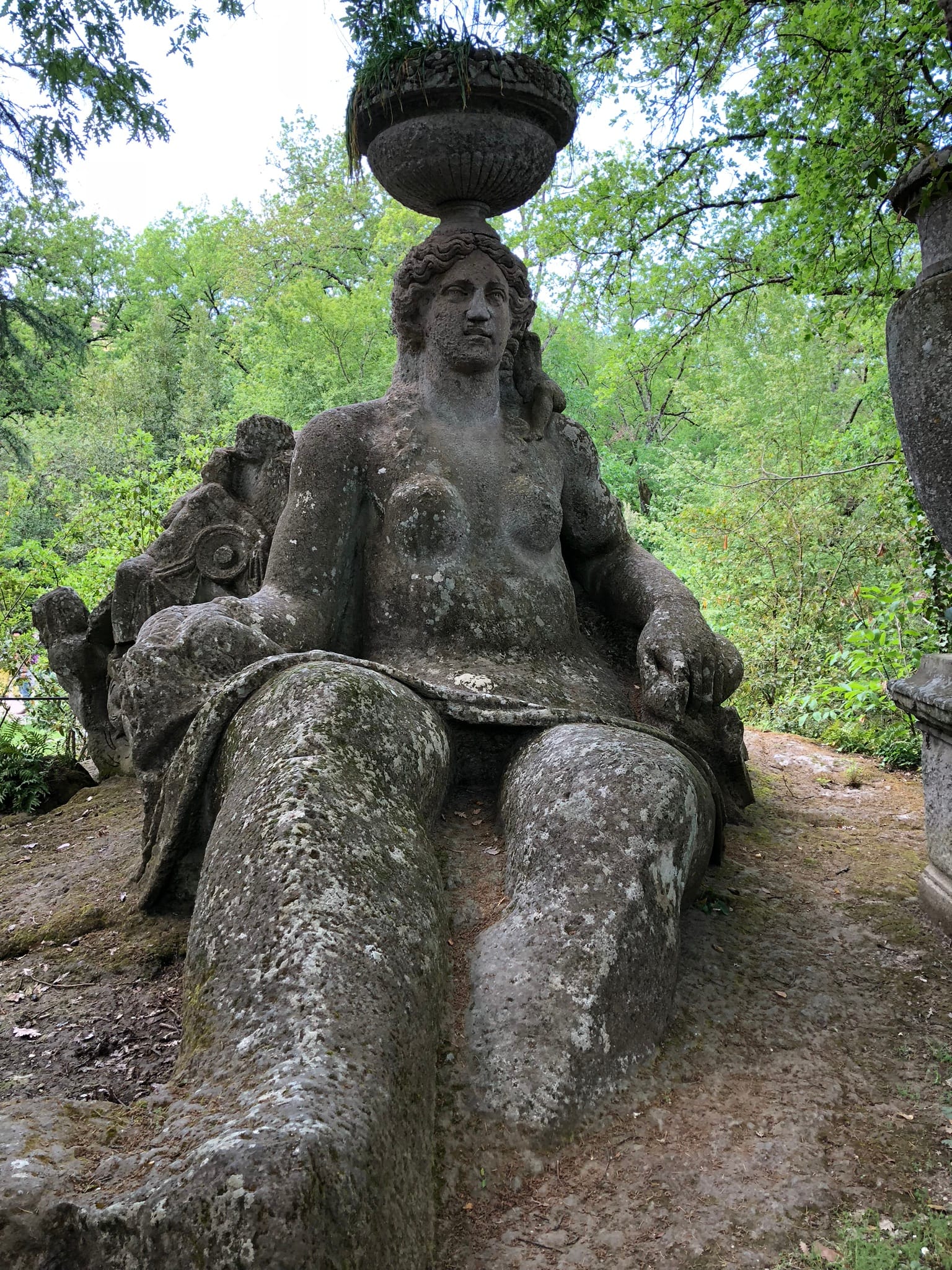 Colossal carved stone statue of Ceres seated on the ground with a large basket of fruit and wheat on her head, surrounded by foliage in the Sacro Bosco of Bomarzo.