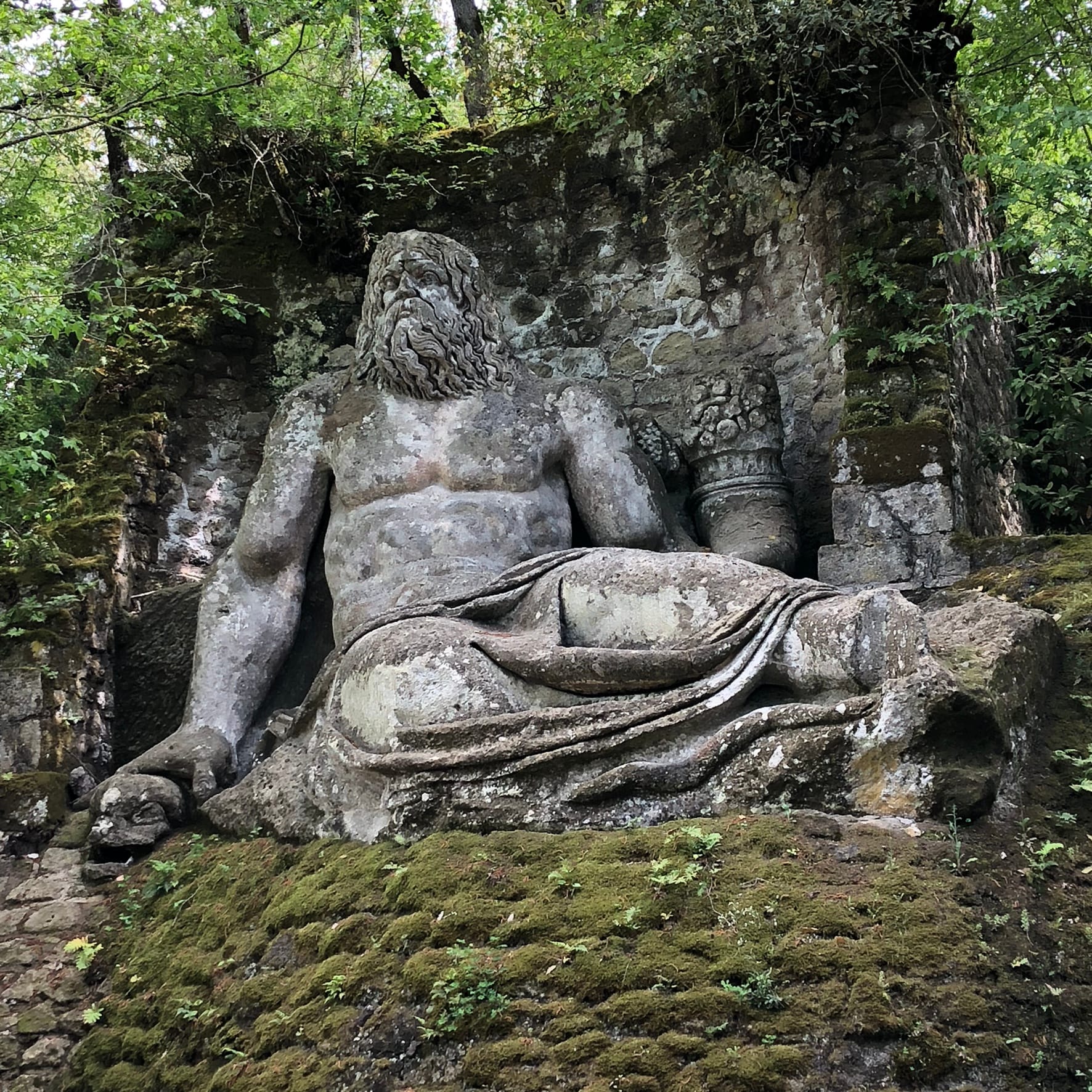 Colossal reclining stone statue of Neptune holding a dolphin in a dry basin among the trees at Bomarzo.