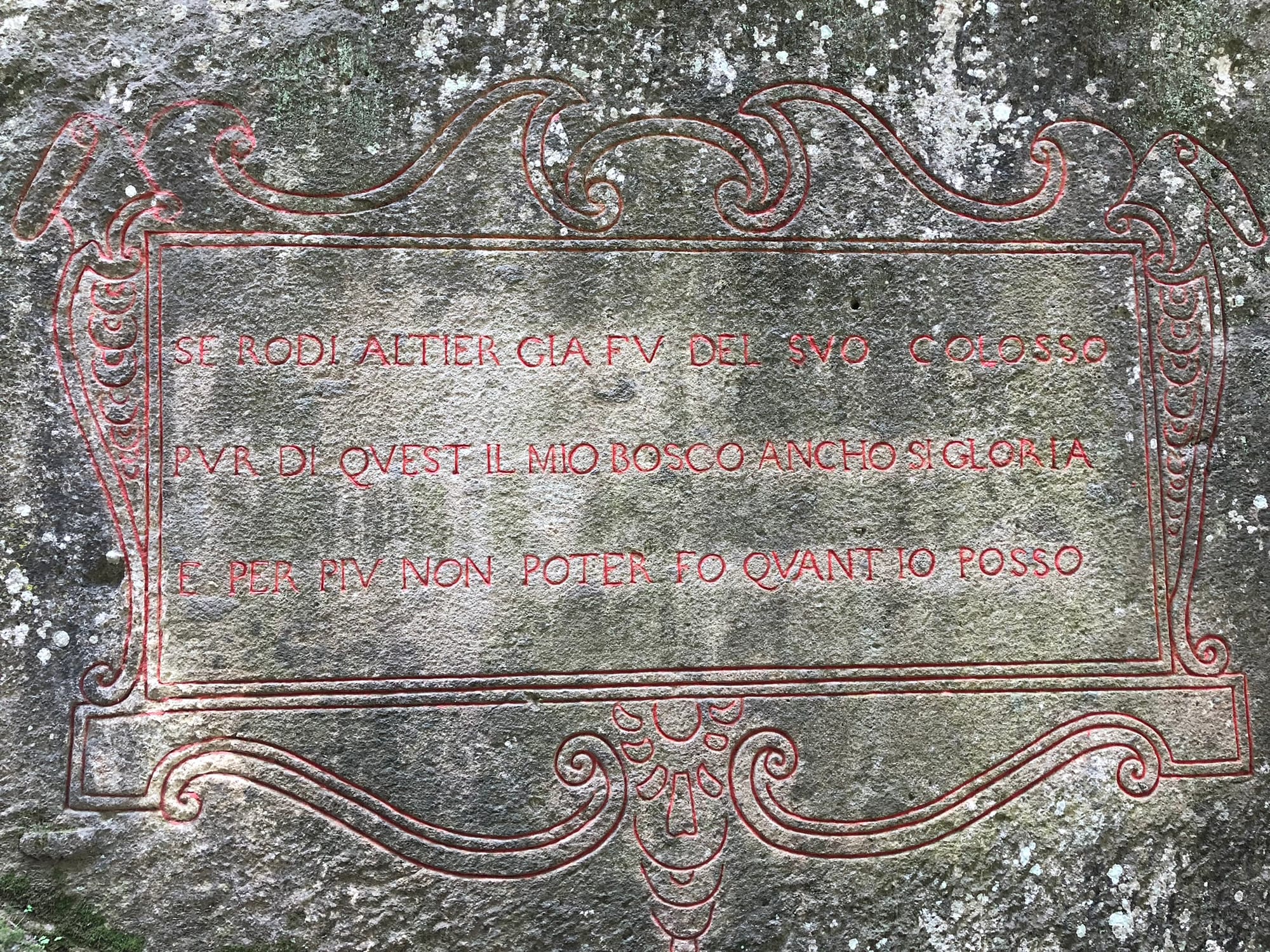 A close-up of a massive, moss-covered peperino stone base next to giant carved legs, featuring the 16th-century Italian inscription "SE RODI ALTIER GIA FV DEL SVO COLOSSO PVR DI QVEST'IL MIO BOSCO ANCHO SI GLORIA E PER PIV NON POTER FO QVANT'IO POSSO" in weathered Roman capital lettering.