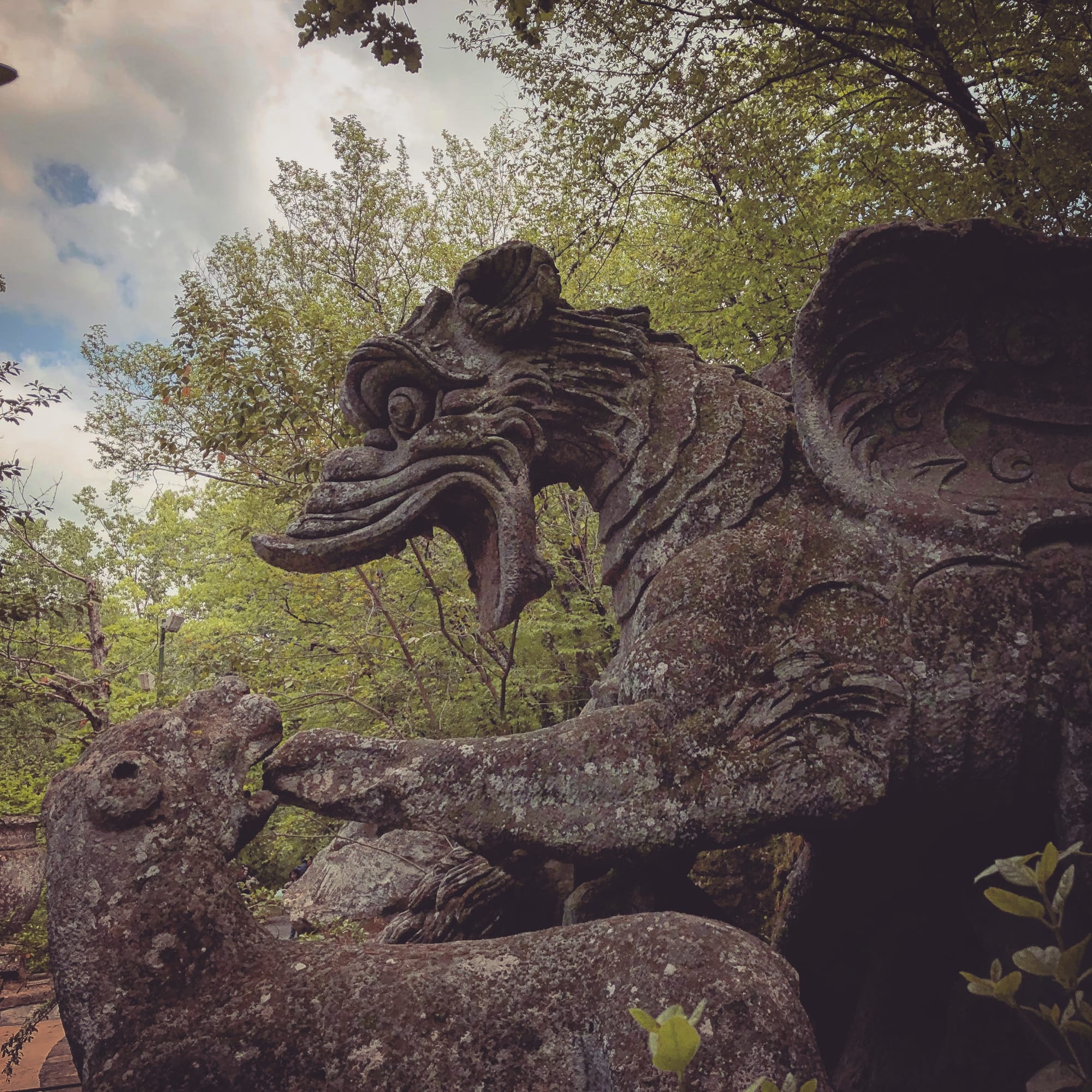 Side view of the stone dragon attacked by lions at Bomarzo, partially framed by moss and trees.