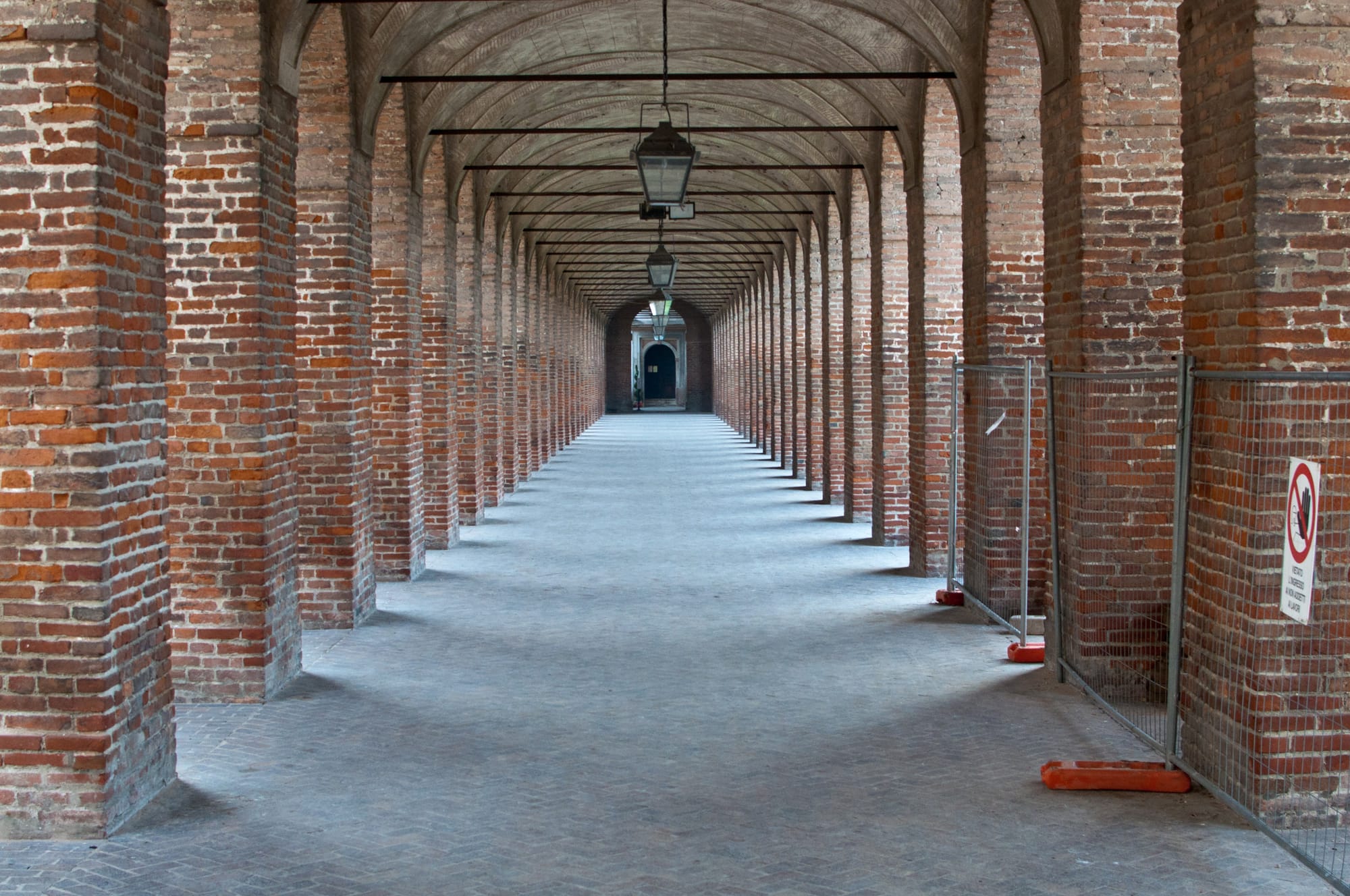 Long brick arcade with a barrel vault and hanging lanterns beneath the Galleria degli Antichi in Sabbioneta, empty to the vanishing point