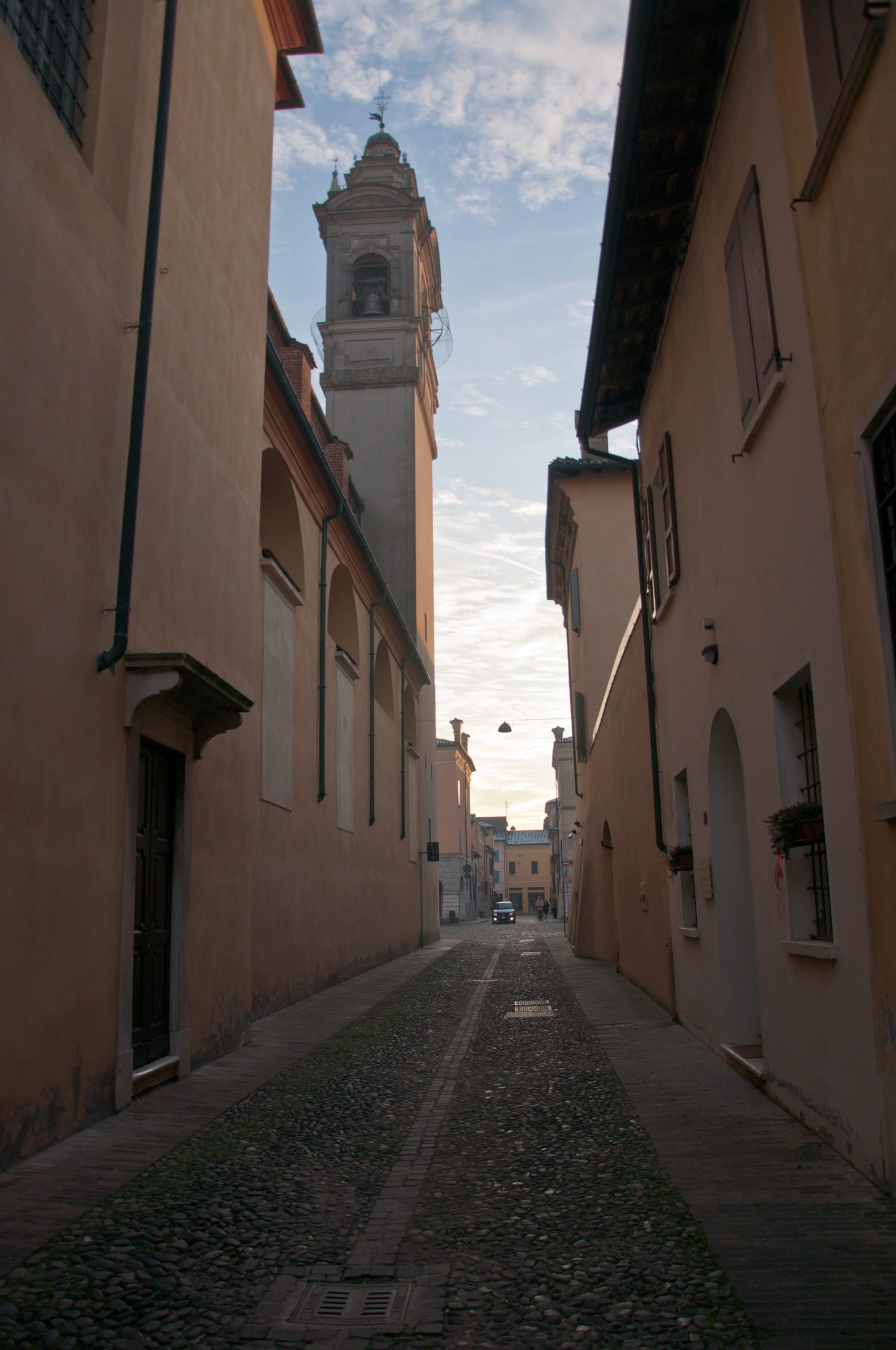 Narrow cobbled street in Sabbioneta between plastered walls, with a baroque bell tower against the sky and a single car in the distance