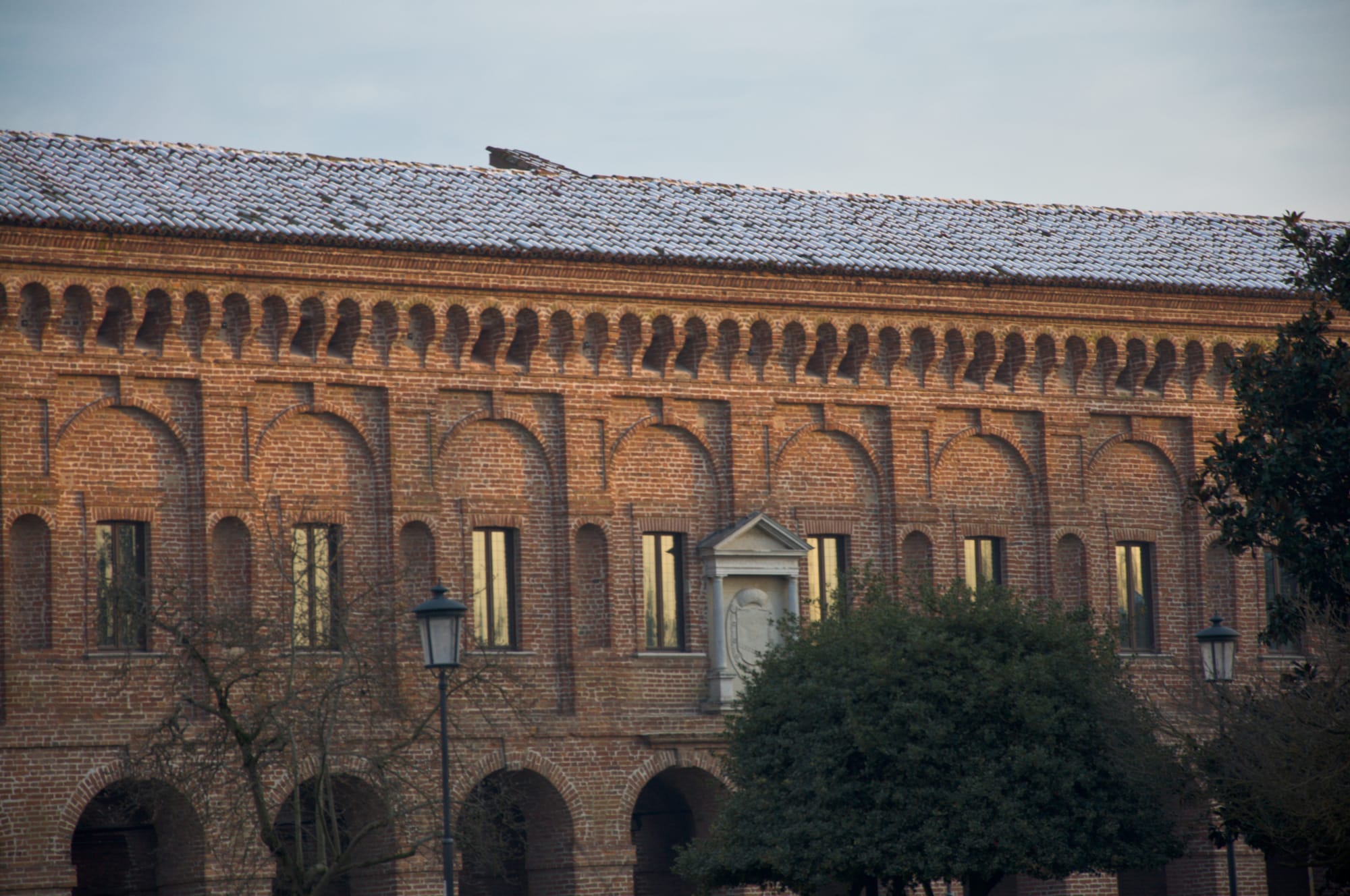 Long brick façade with arched niches and an arcade, its tiled roof lightly covered in snow, behind bare winter trees