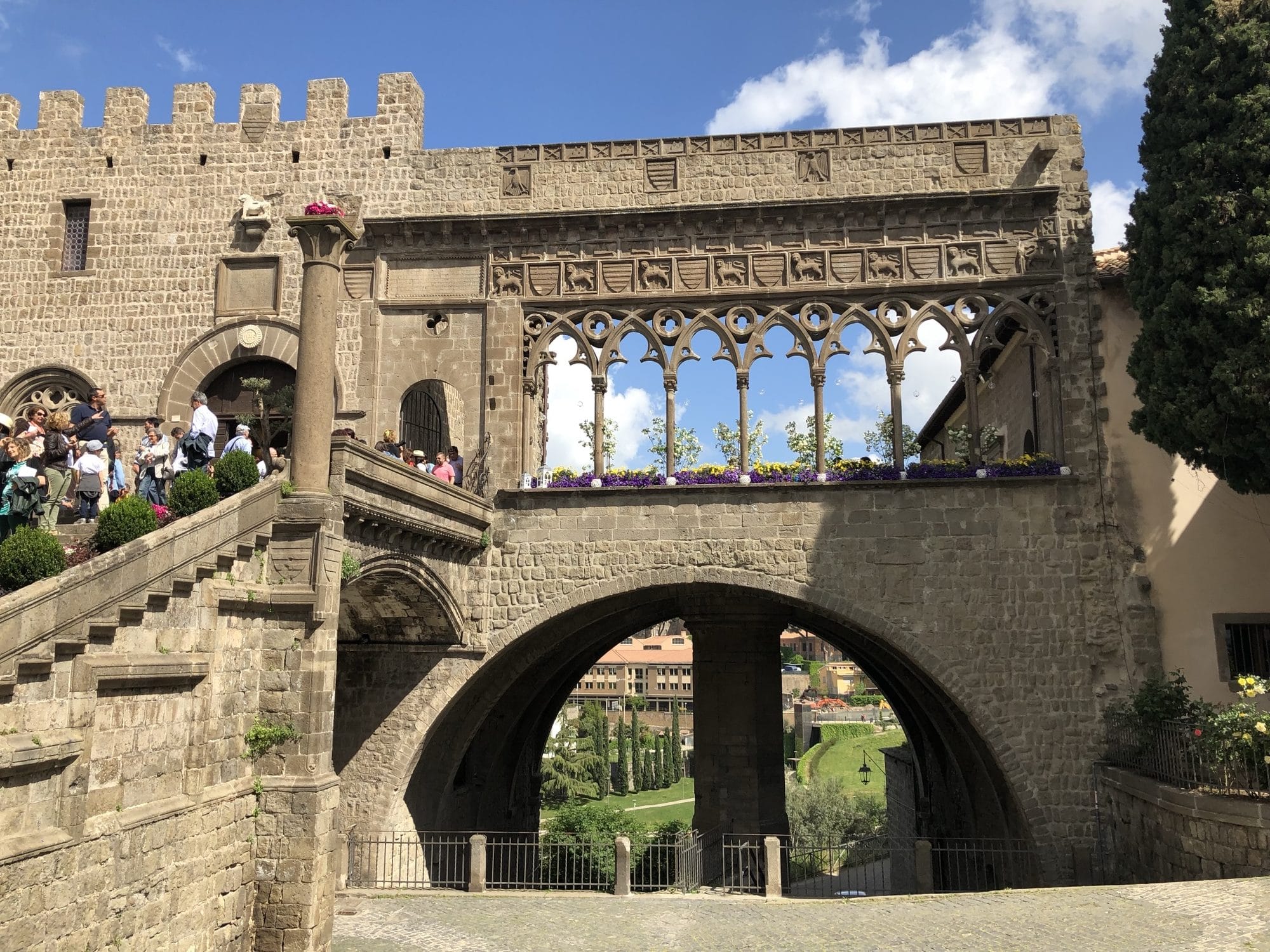 The Gothic loggia of the Palazzo dei Papi in Viterbo, its traceried arches decorated with flowers, and the crowded staircase below, under a spring sky