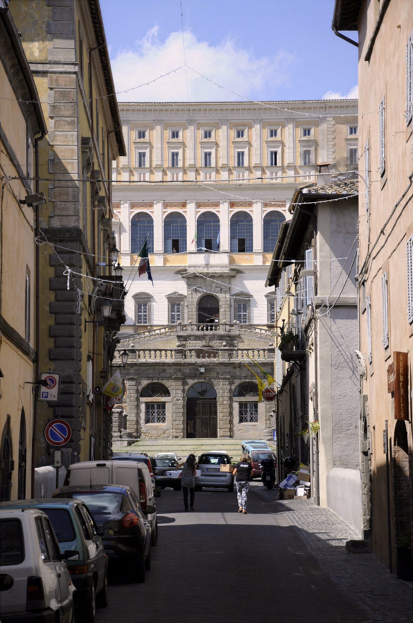 Looking up the last stretch of the Via Dritta in Caprarola: the façade and loggia of Villa Farnese fill the end of the narrow straight street, with cars parked on the left
