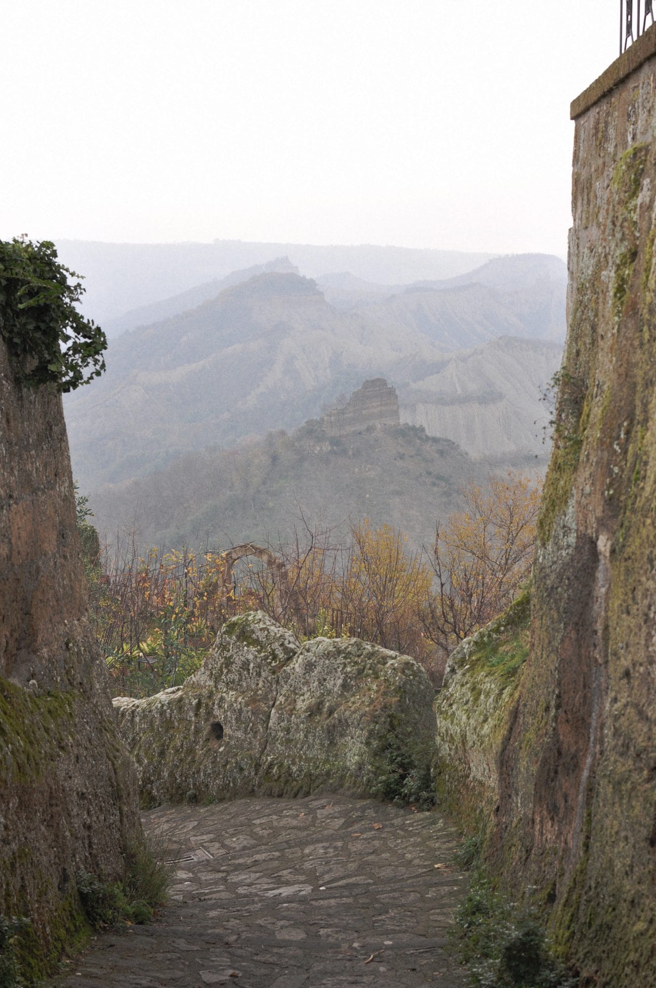 A narrow lane in Civita di Bagnoregio between two walls ending at an iron railing, with the calanchi valley beyond in winter haze
