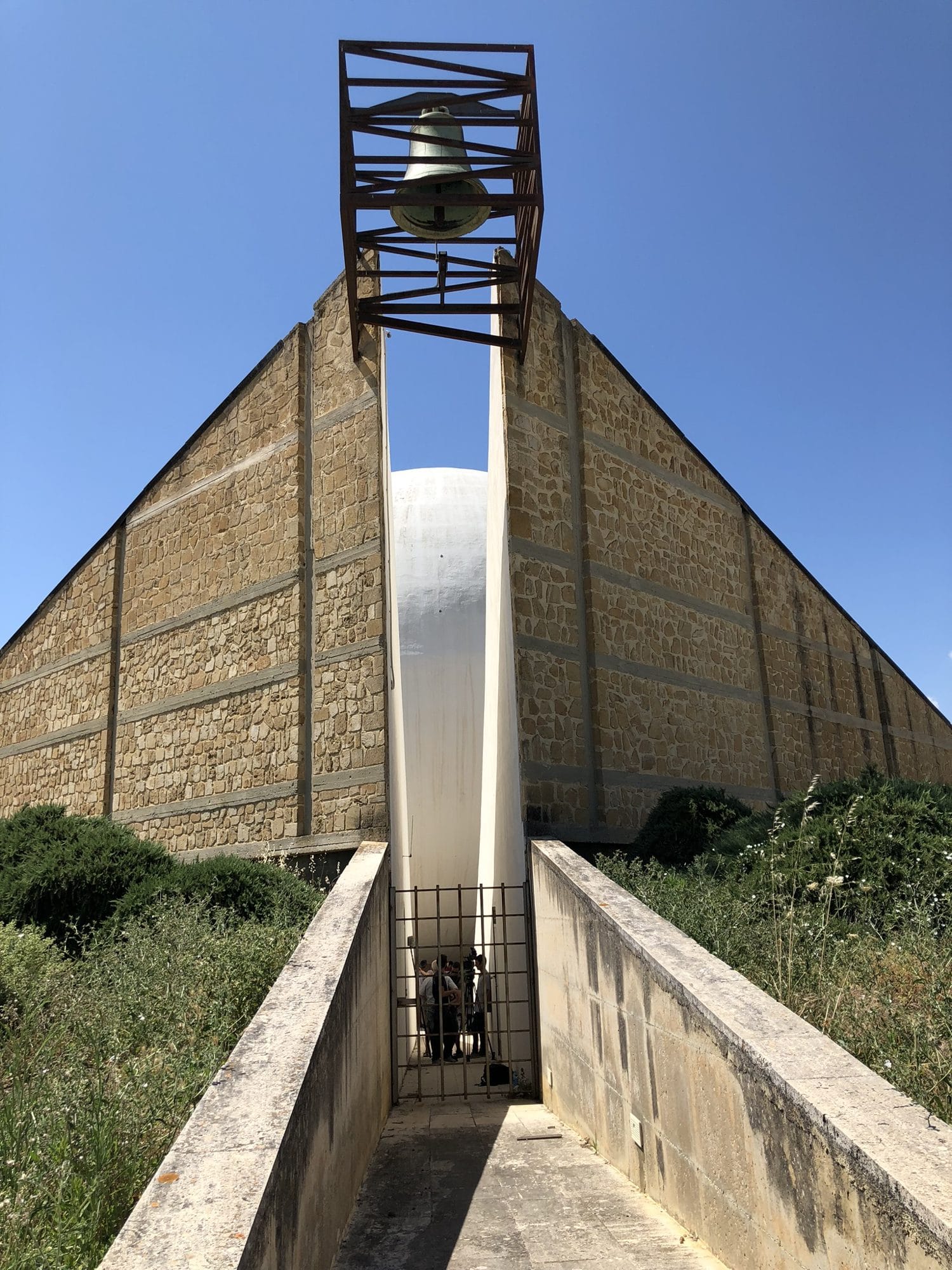A tall open bell frame above a slot between two sloping stone walls of the Chiesa Madre of Gibellina Nuova