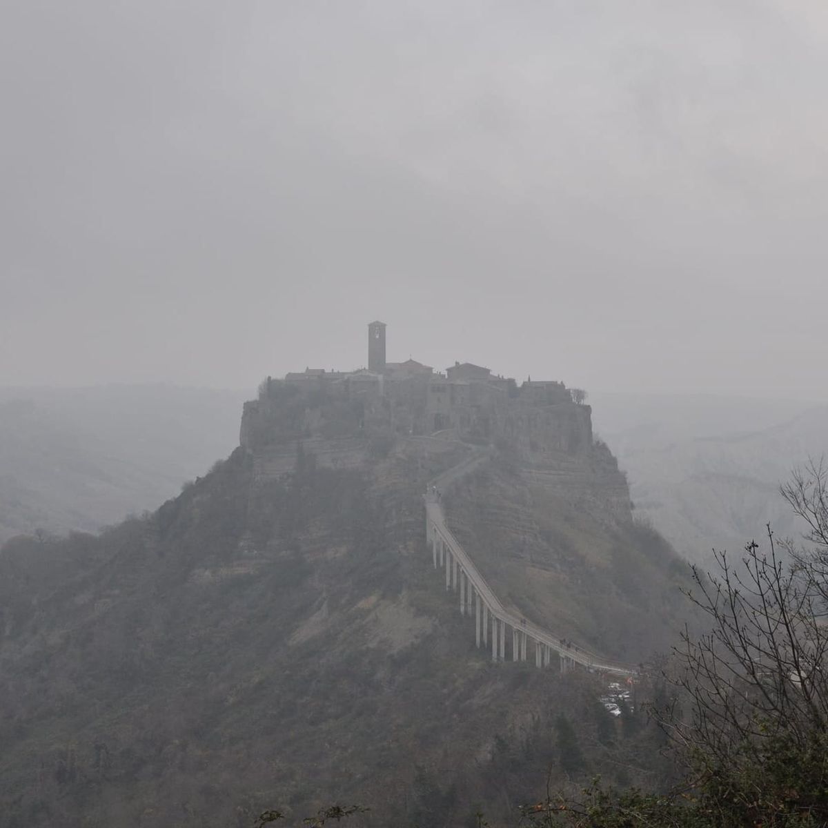 A wide landscape shot of the hilltop town of Civita di Bagnoregio rising like an island out of dense grey fog, connected by a long, steep concrete footbridge.