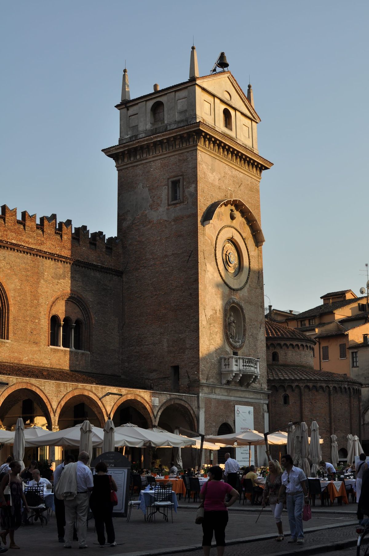 The Torre dell'Orologio in Mantua in late golden light, its gilded astronomical dial set into the brick, with the crowded restaurant tables of Piazza delle Erbe beneath it.