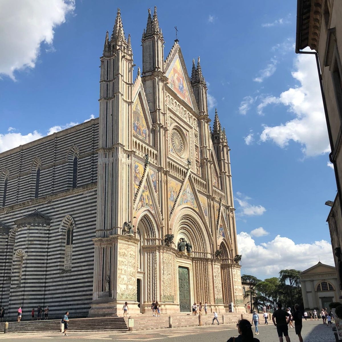 Oblique view of Orvieto Cathedral under a blue sky, showing the intersection where the mosaic-covered Gothic front screen meets the black-and-white striped side wall.