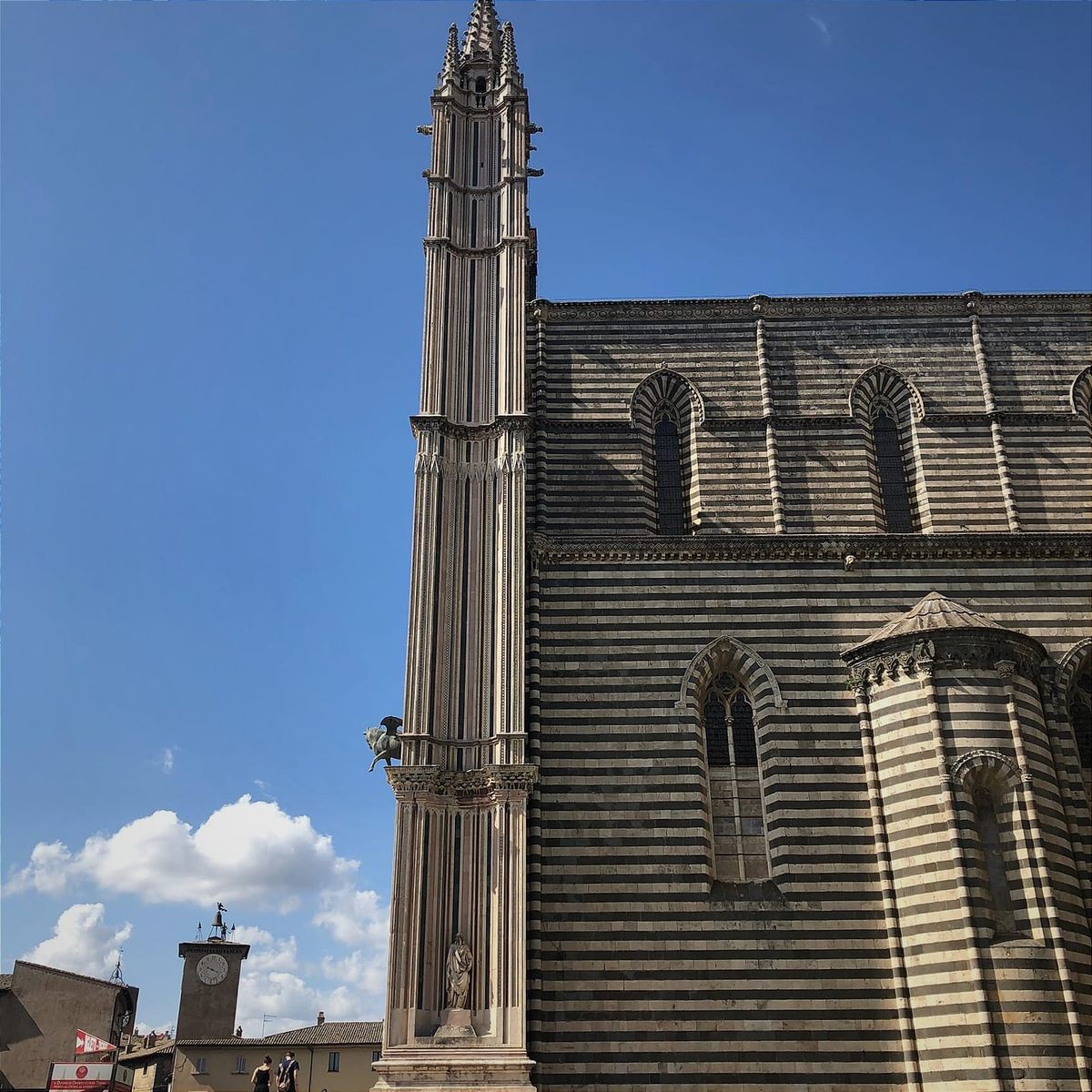 Orvieto Cathedral side view: Distinctive zebra-striped marble wall with tall ornate Gothic spire, arched windows, and distant clock tower against blue sky.