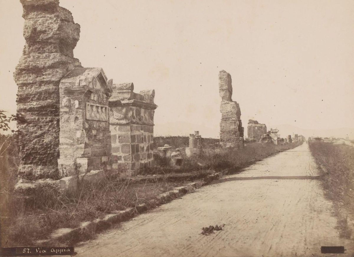Sepia photograph of a straight, empty country road lined with ruined tombs and brick walls with marble reliefs set into them, with no trees along it