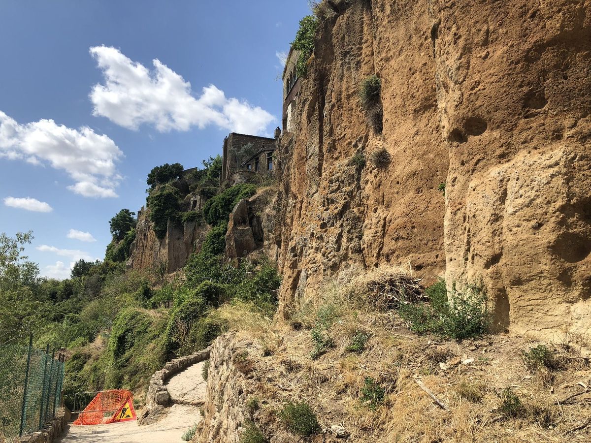 A footpath beneath the eroded tufa cliff of Civita di Bagnoregio, under a blue summer sky