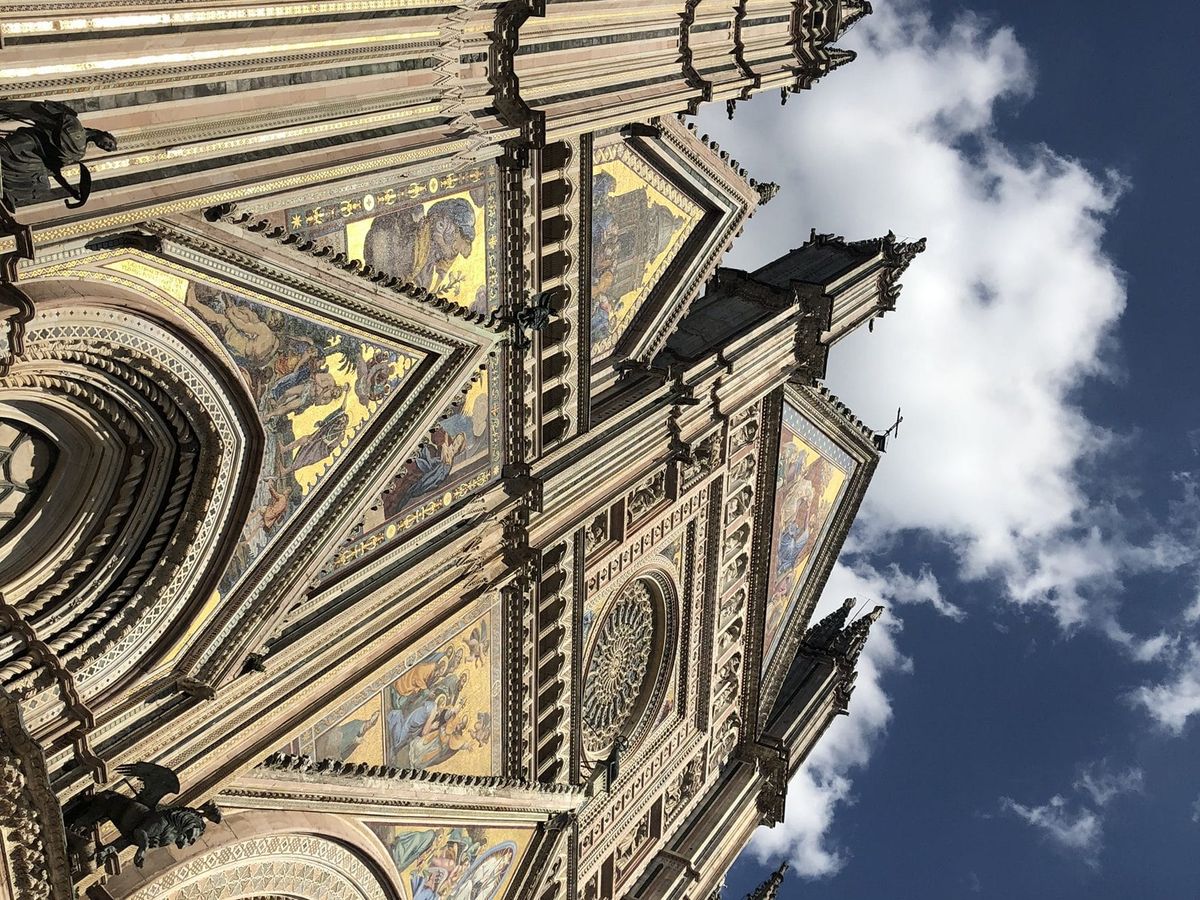 Looking up at the Gothic façade of Orvieto Cathedral: gold mosaics, carved pinnacles and the rose window against a blue sky with white clouds