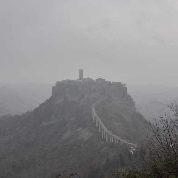 A wide landscape shot of the hilltop town of Civita di Bagnoregio rising like an island out of dense grey fog, connected by a long, steep concrete footbridge.