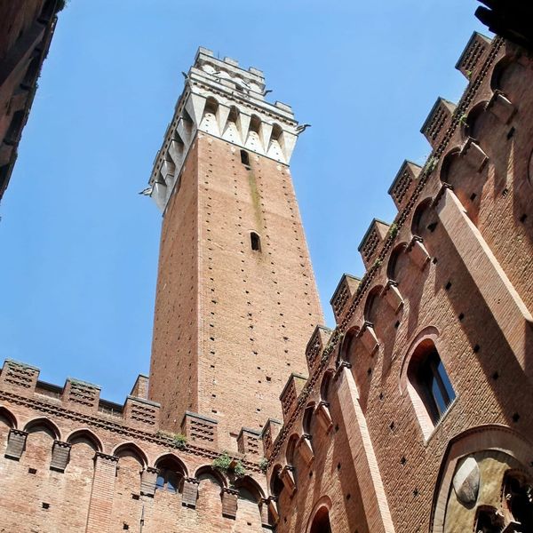 A dramatic low-angle shot looking straight up the vertical brick shaft of the Torre del Mangia toward its white stone battlements against the sky.