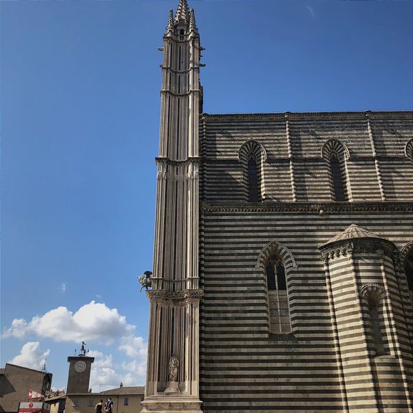 Orvieto Cathedral side view: Distinctive zebra-striped marble wall with tall ornate Gothic spire, arched windows, and distant clock tower against blue sky.