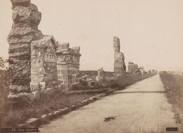 Sepia photograph of a straight, empty country road lined with ruined tombs and brick walls with marble reliefs set into them, with no trees along it