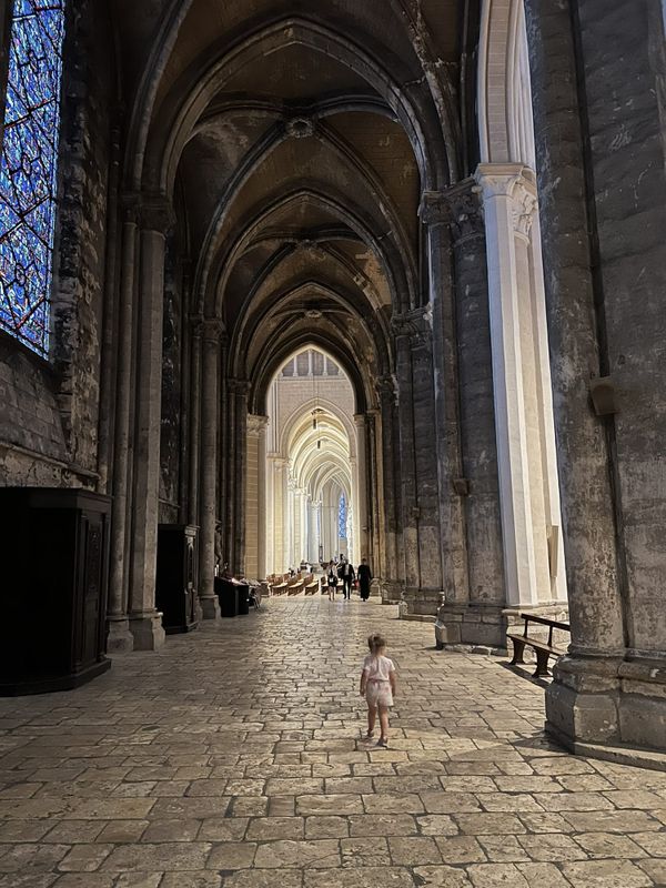 A small child walks away down a side aisle of Chartres Cathedral, between blackened vaults and a half-cleaned pier, towards the bright nave at the far end