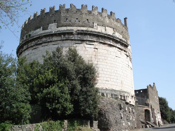The round travertine tomb of Caecilia Metella crowned with medieval battlements, rising above holm oaks beside the Appian Way under a clear blue sky