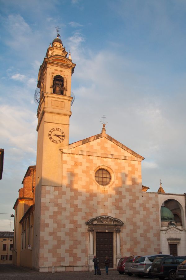 The chequered pink and white façade of Santa Maria Assunta in Sabbioneta lit by a winter sunset, with parked cars in the square