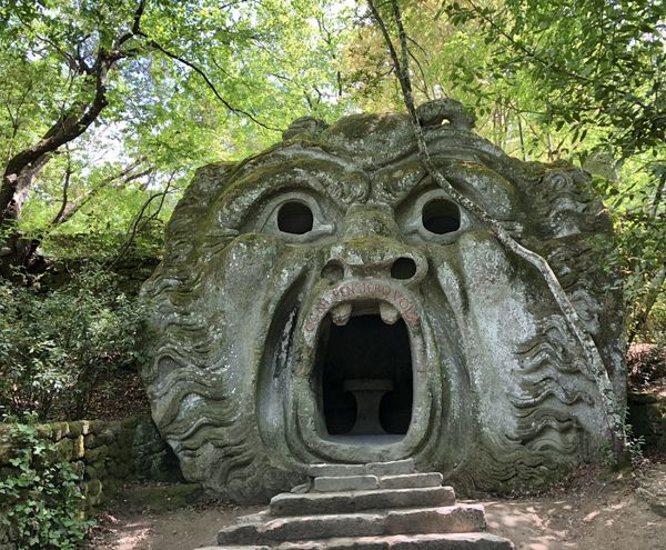 The giant stone head of the Orcus at the Monster Park of Bomarzo, its wide-open mouth a doorway with steps leading in, among the trees